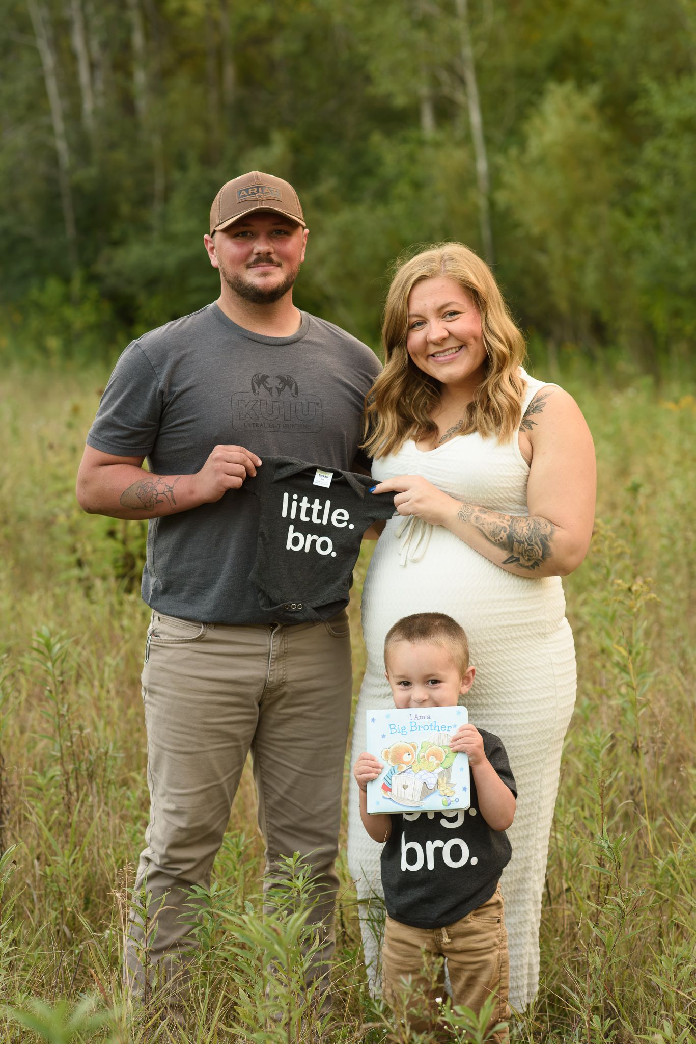 Couple holding little bro shirt with toddler in big bro shirt announcing pregnancy outdoors.
