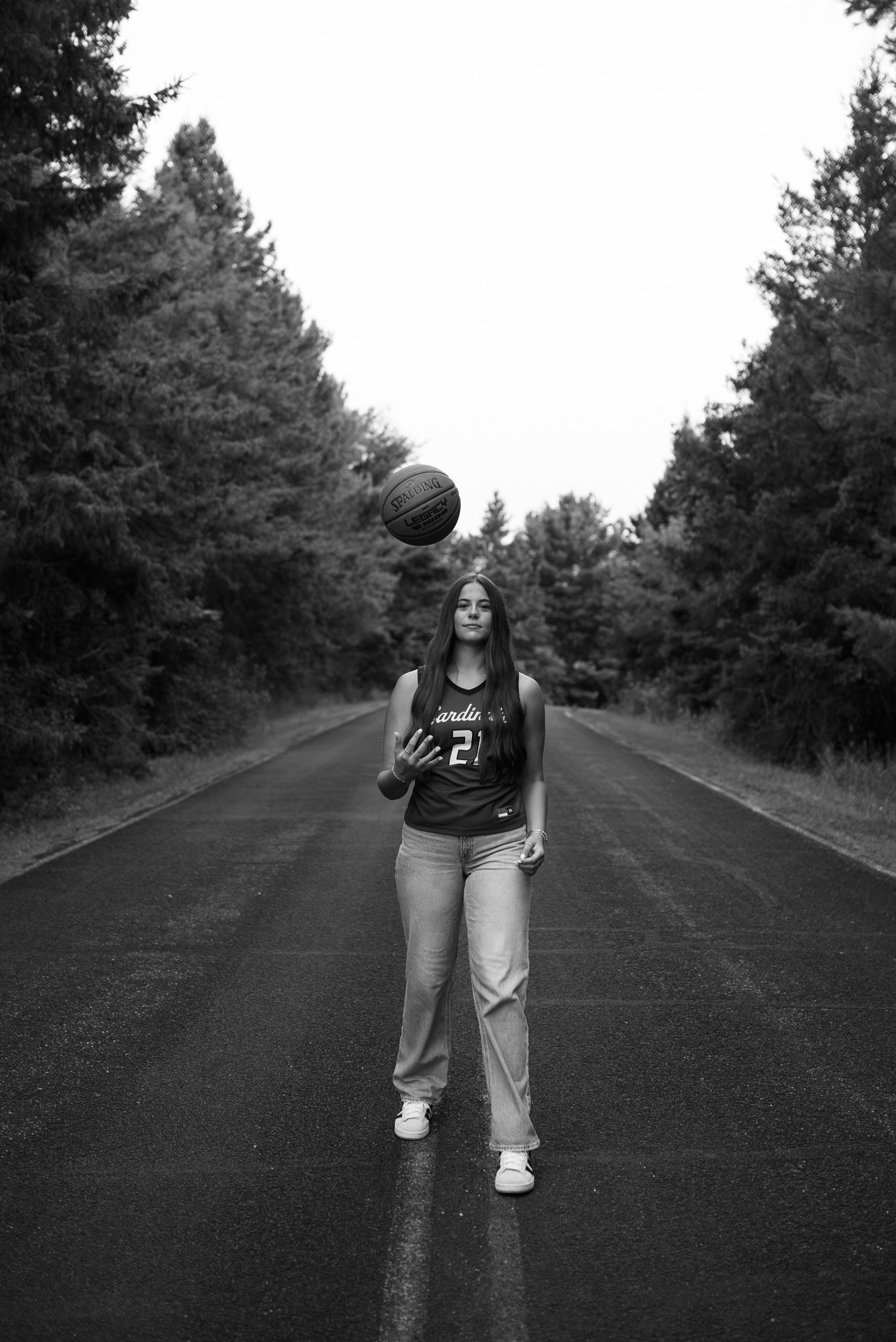 Black and white photo of a teen girl in basketball jersey tossing a ball on a tree-lined road.