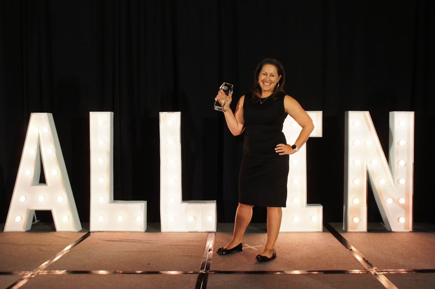 Woman in black dress holds award trophy in front of illuminated ALL IN letter signs on stage.