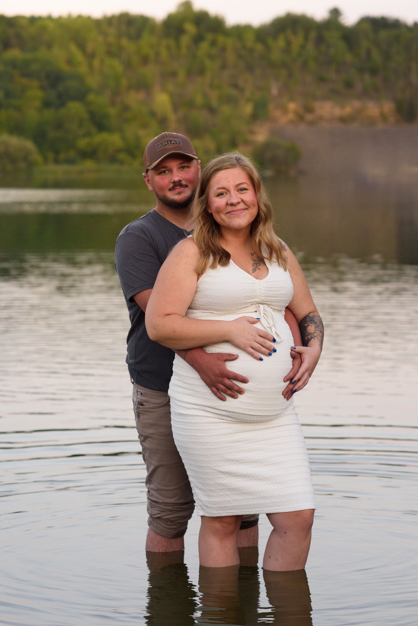 Couple standing in lake water embracing pregnant belly at sunset.