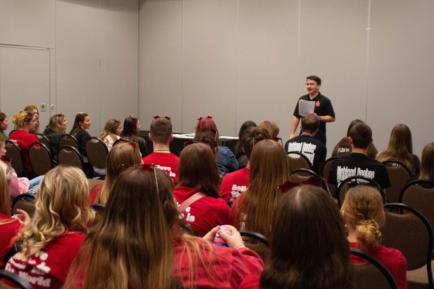 A presenter in black addresses a room of students in red shirts at a conference.