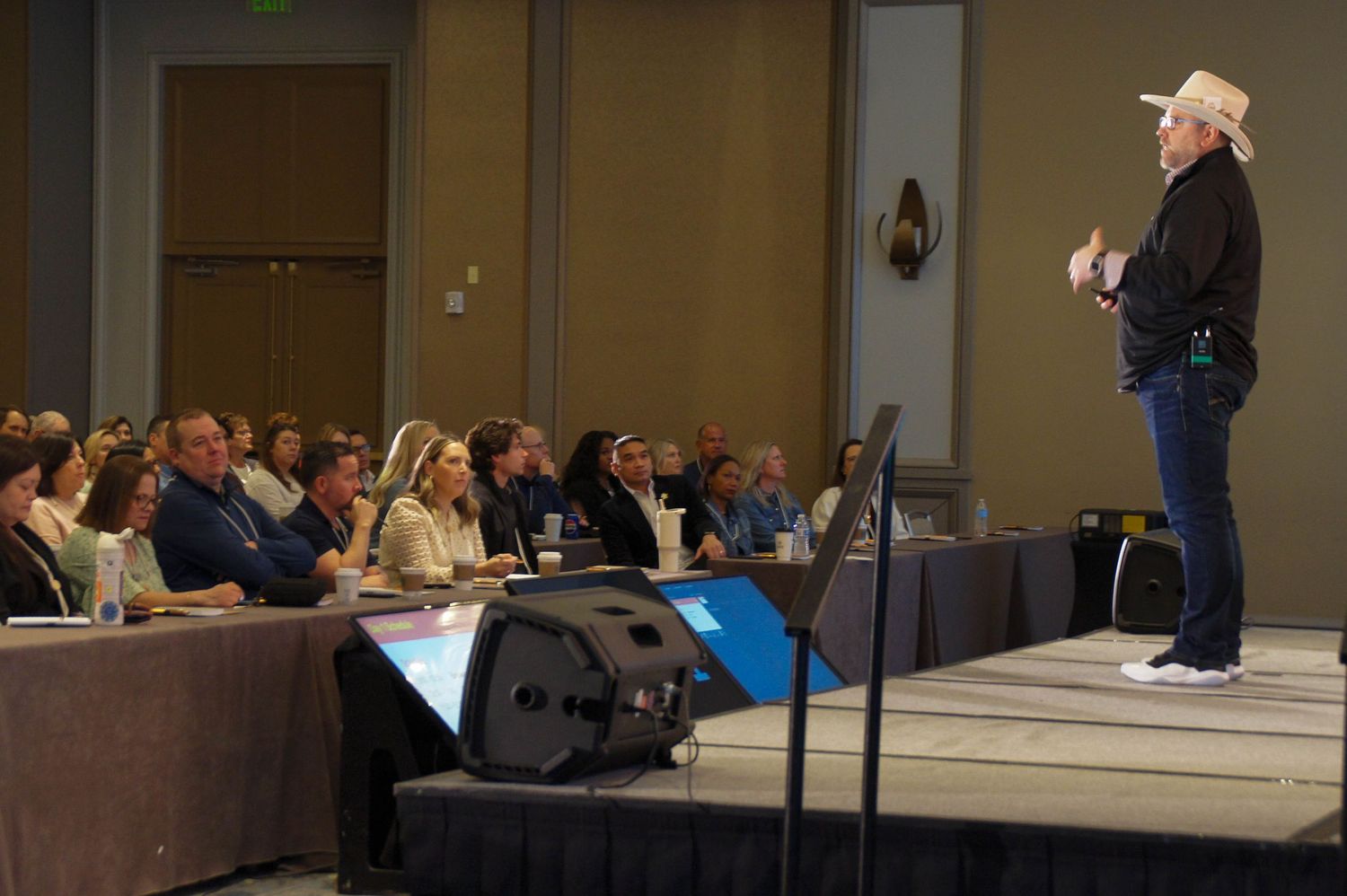 A speaker in a cowboy hat addresses a large audience at a professional conference event in a hotel ballroom.