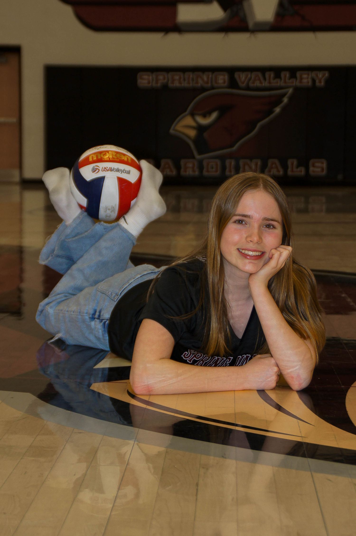 Smiling girl in Spring Valley volleyball shirt lying on gym floor with volleyball.