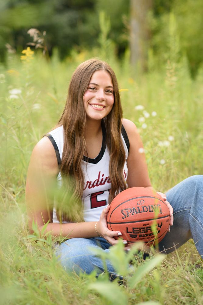 Smiling teen girl in Cardinals jersey holds Spalding basketball while sitting in green meadow for senior portrait.