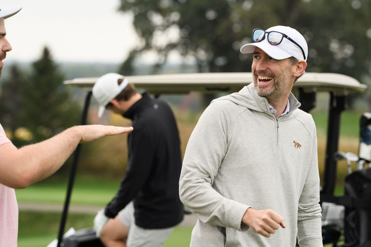 Two men laughing on a golf course near a golf cart on a cloudy day, wearing casual golf attire.
