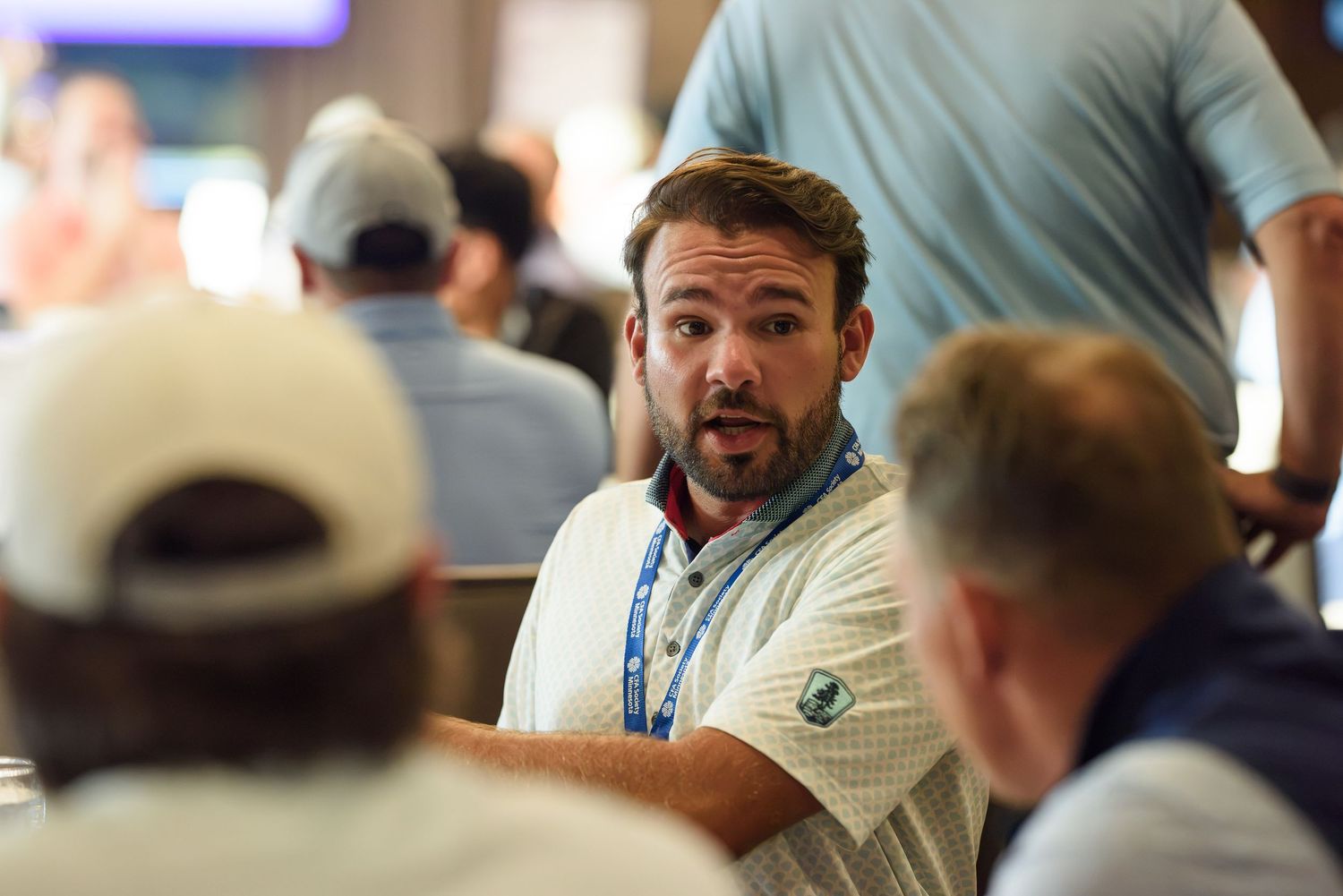 Man in white polo with blue lanyard talking to others at an indoor golf event.