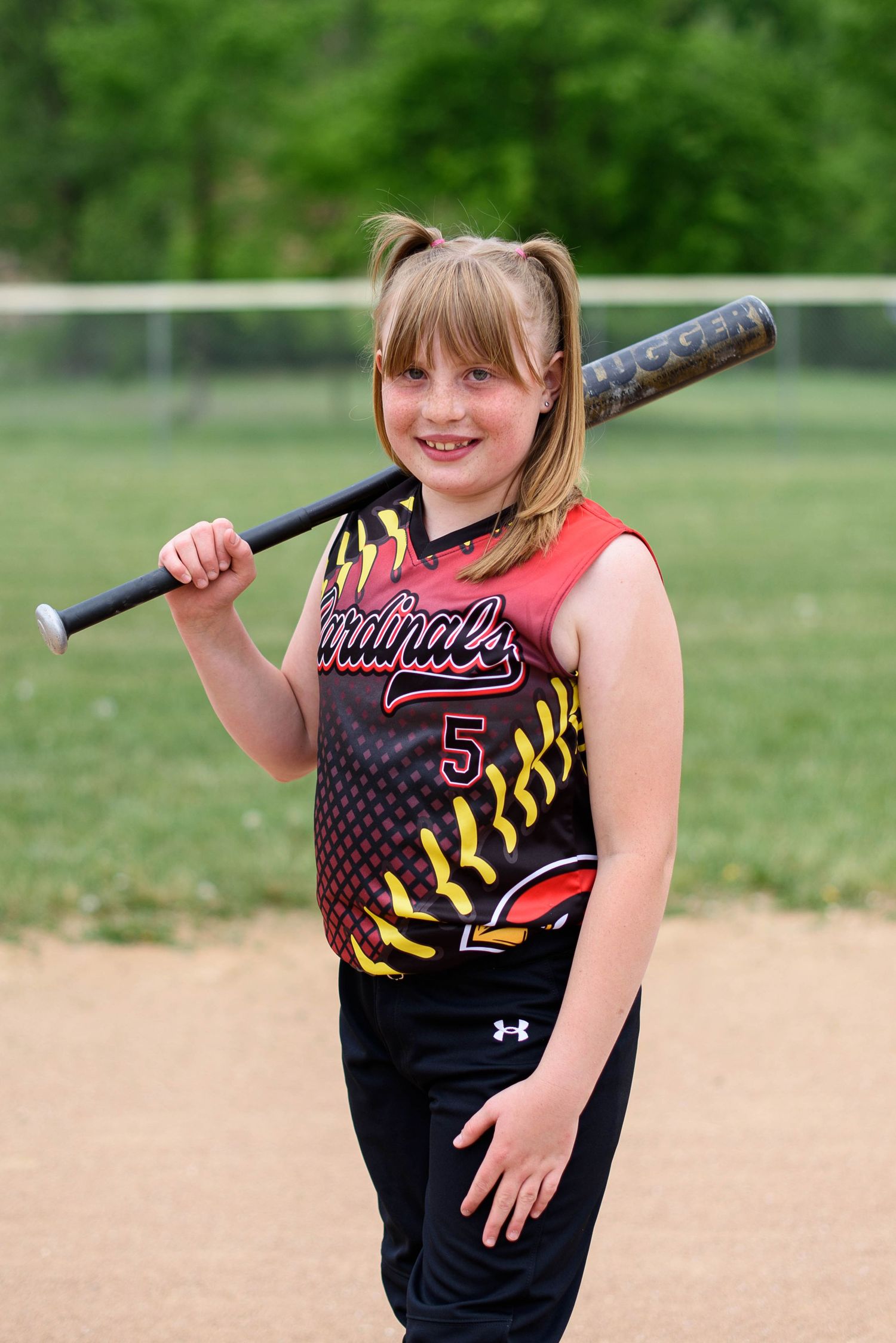 Young Cardinals softball player #5 smiling and posing with a bat resting on her shoulder on a dirt field.