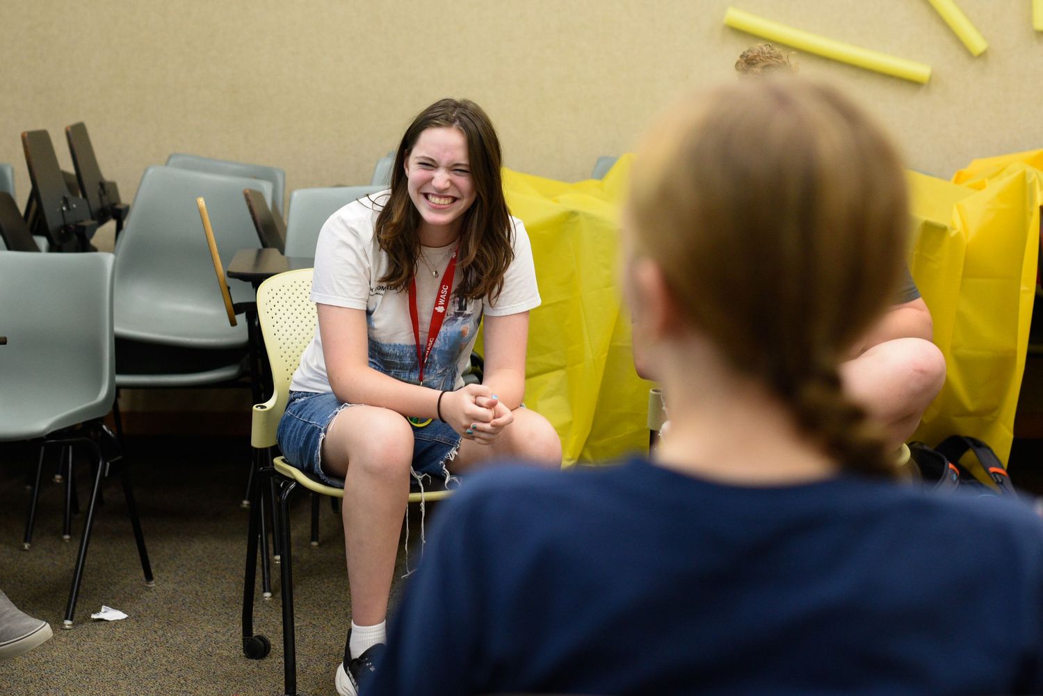 Two young women sitting and smiling during an indoor activity session with yellow chairs in background.