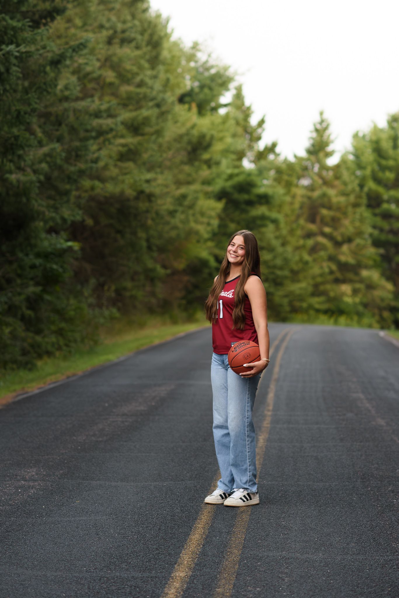 Teen girl in maroon jersey holds basketball while standing on a tree-lined road in jeans and Adidas sneakers.