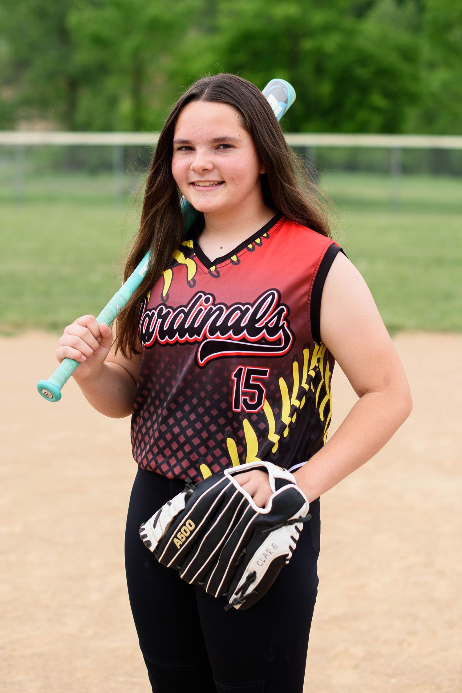 Teen girl in Cardinals #15 softball uniform posing with bat and glove on field.