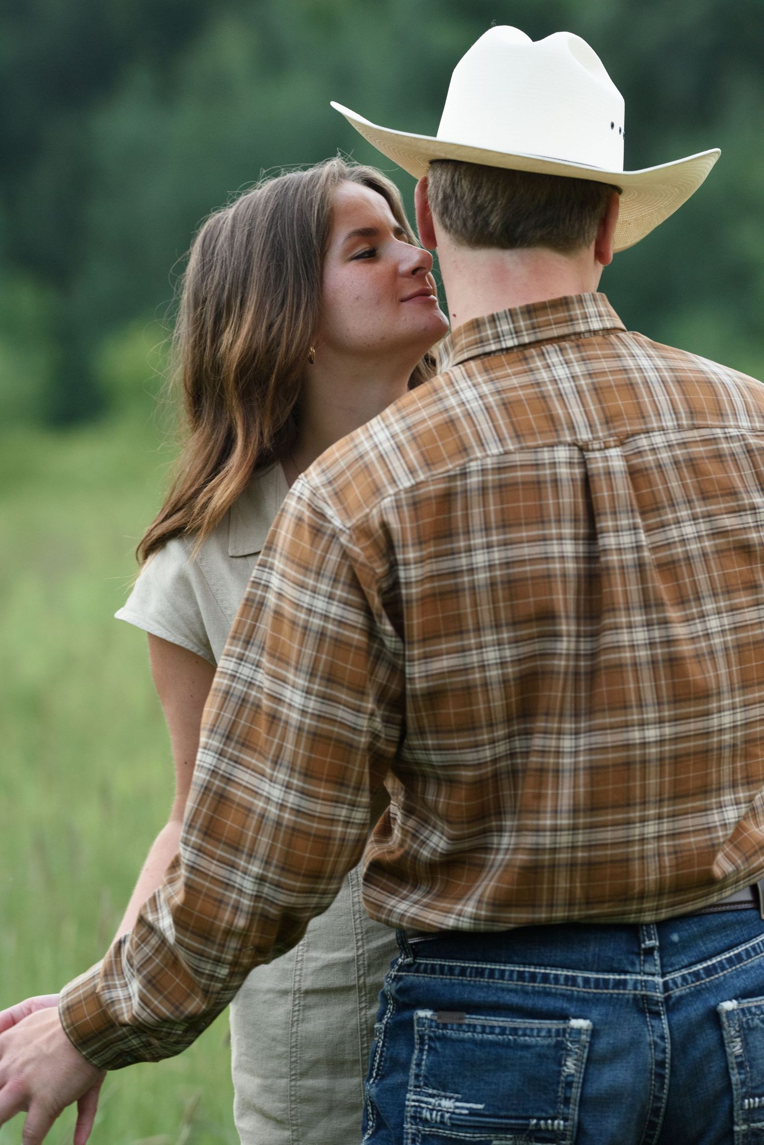 A woman leans in to kiss a man wearing a white cowboy hat and plaid shirt in a green field.