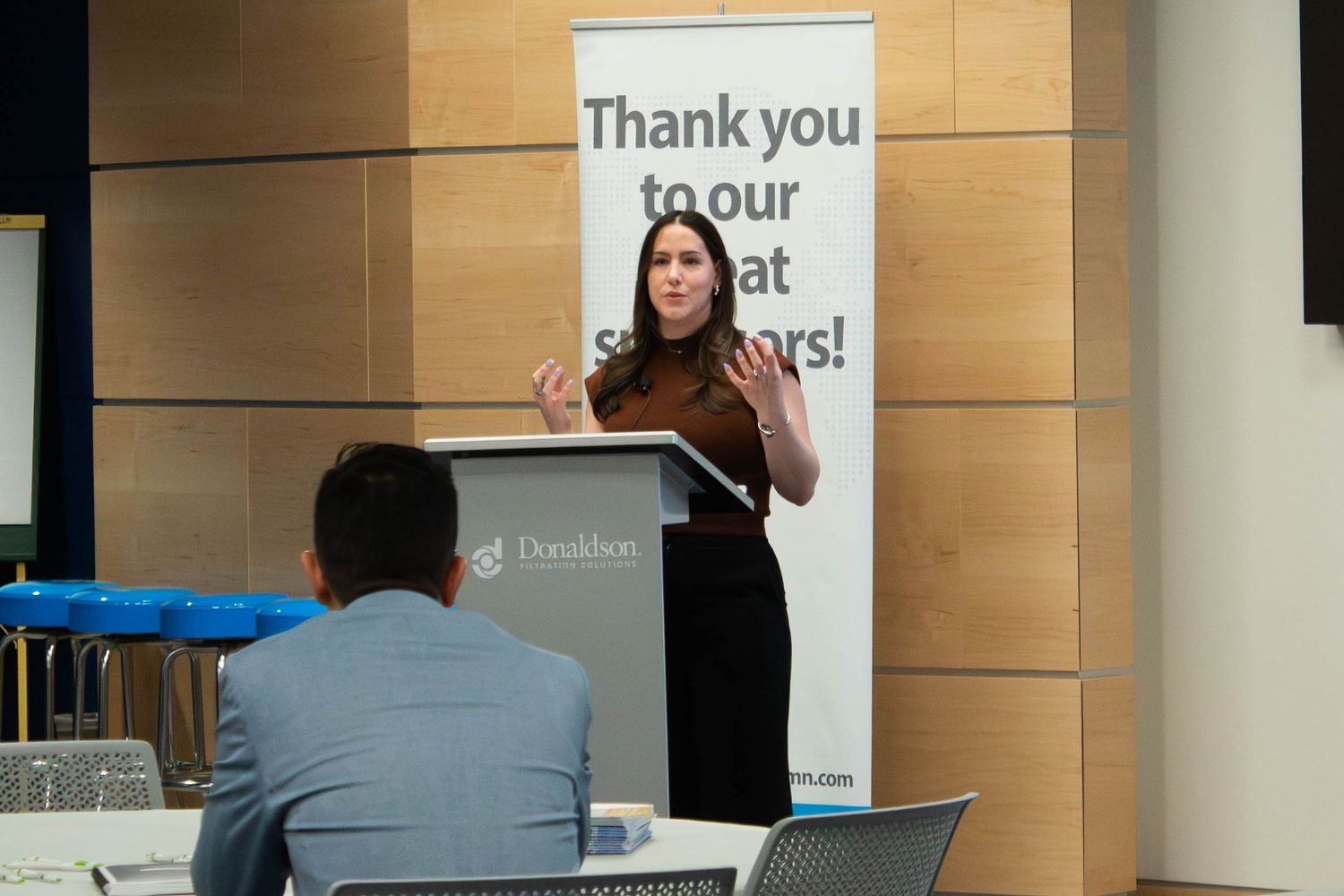 Woman speaking at podium during corporate event with Thank You to our Sponsors banner in background.