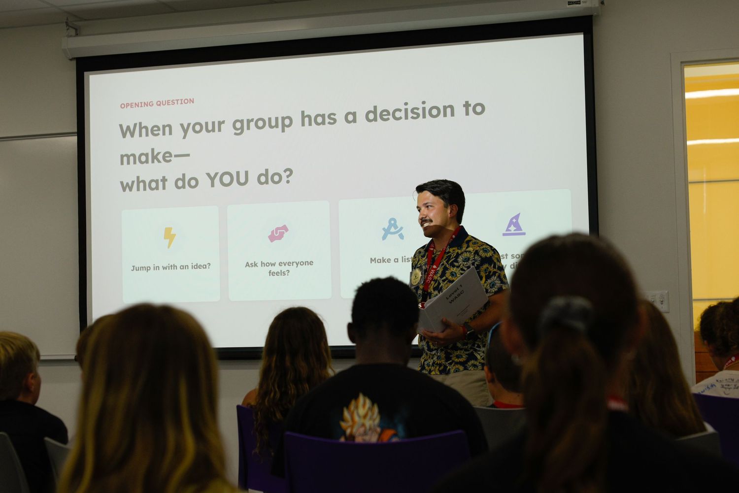 A presenter stands before a projected slide about group decision-making in a modern classroom with seated attendees.