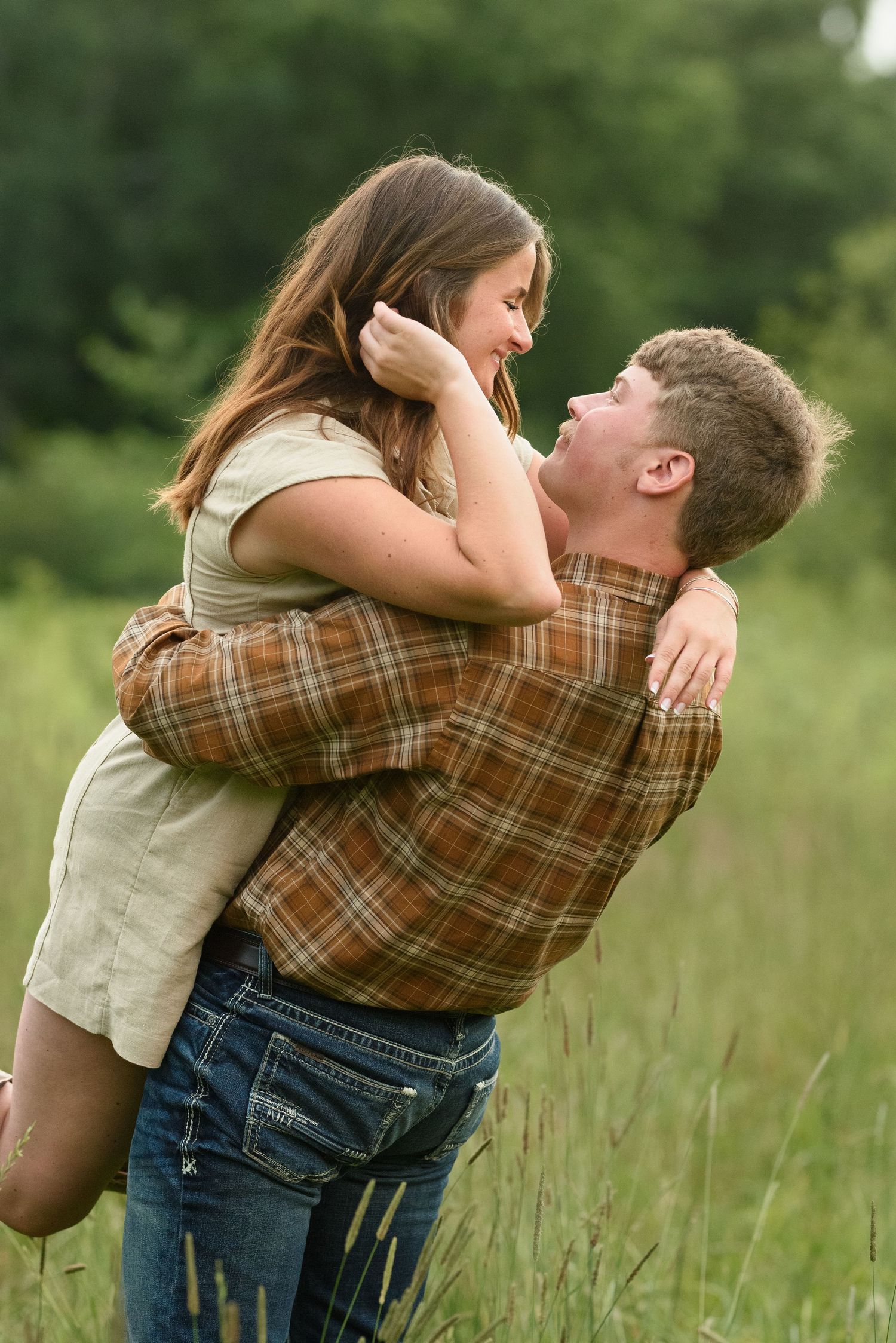 Man lifting woman in his arms laughing in tall green grass field, wearing casual plaid shirt.