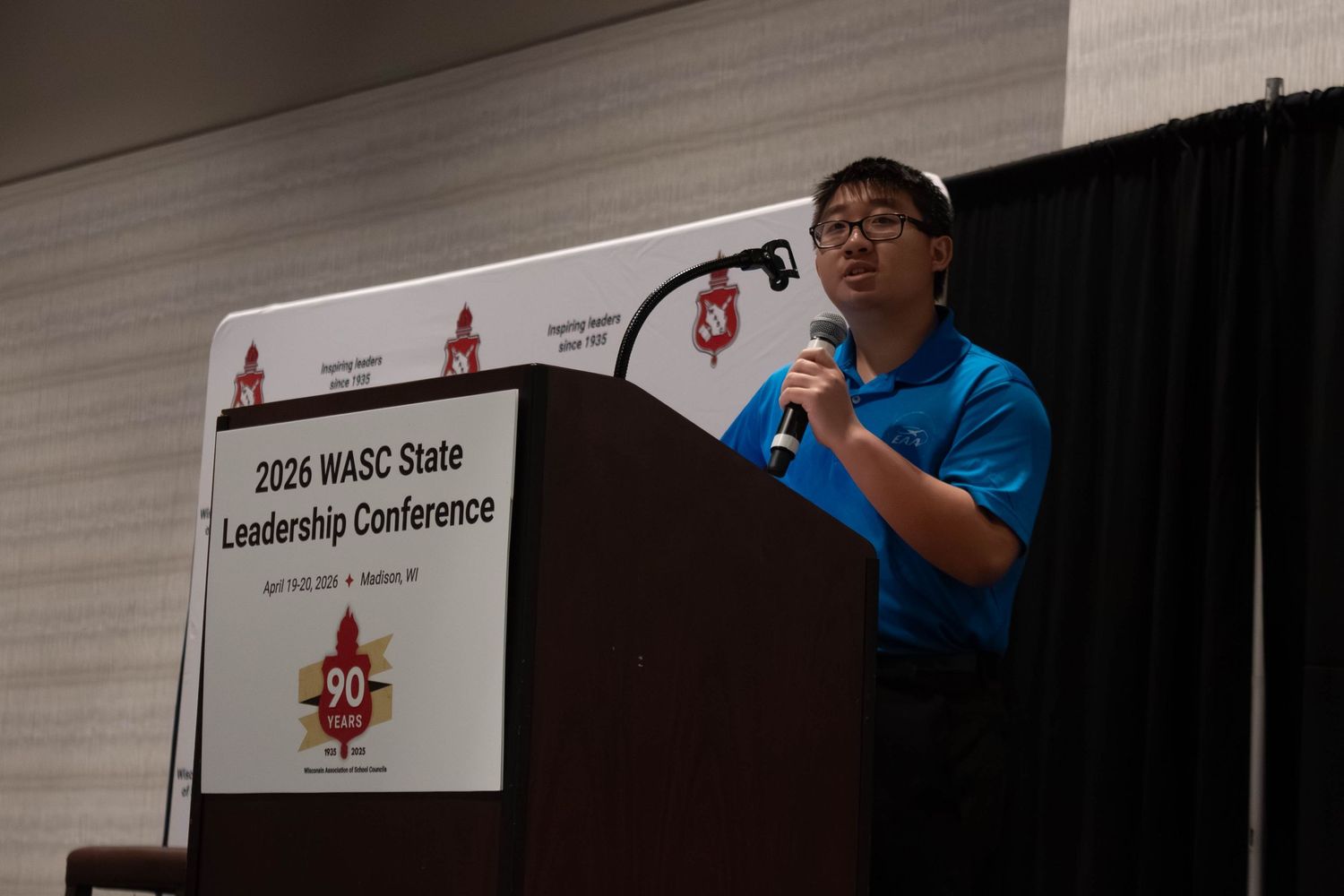 A young man in a blue polo speaks at a podium at the 2026 WASC State Leadership Conference.