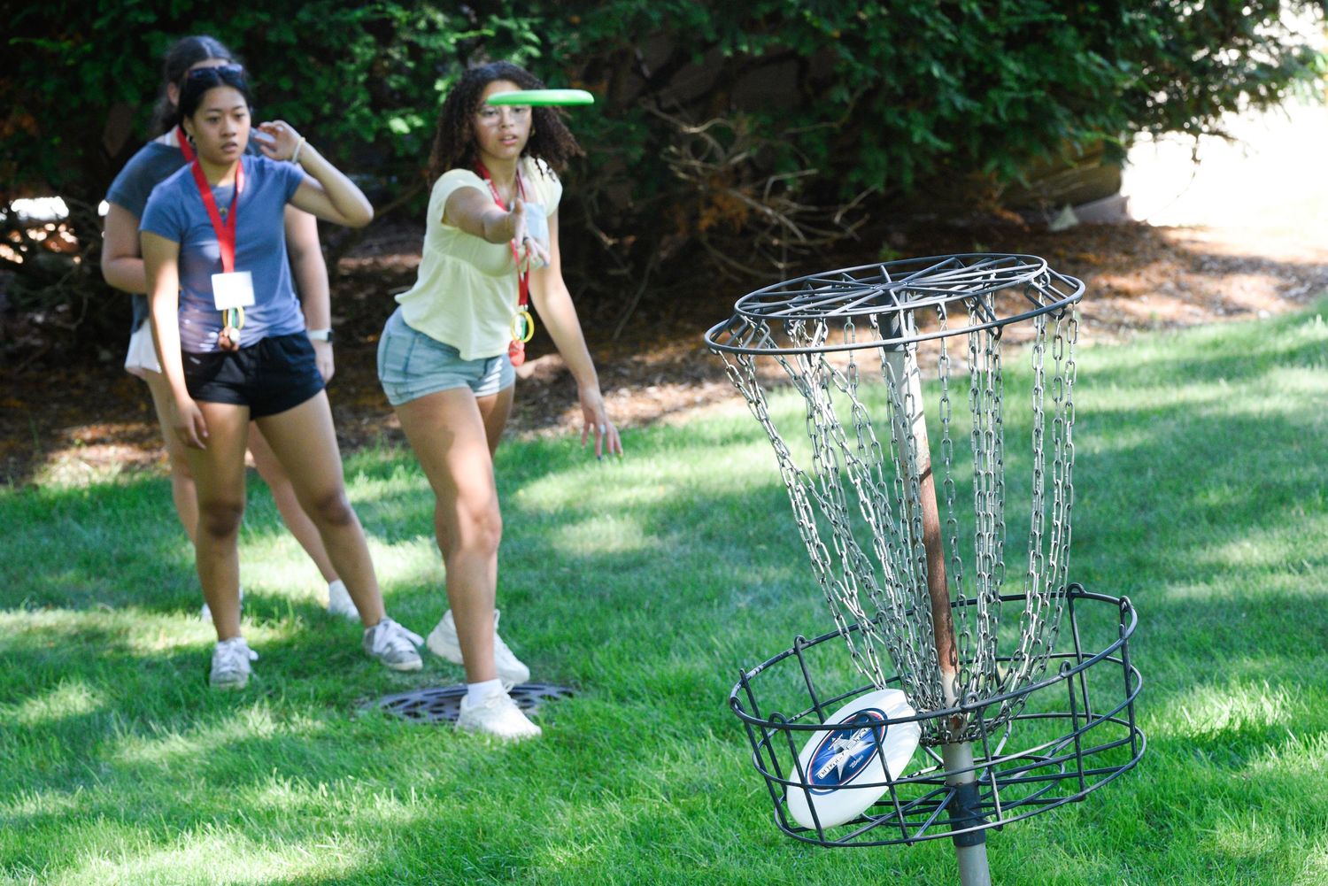 Two young women playing disc golf, one throwing a frisbee toward a metal chain basket on a grassy lawn.