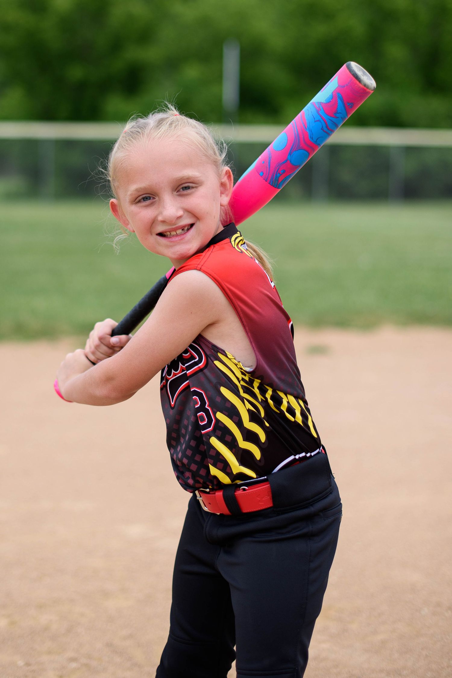 Young girl in softball uniform smiling and posing with a colorful pink bat on a baseball diamond.