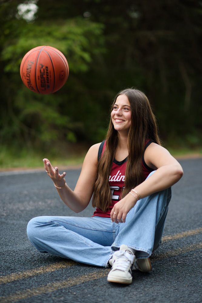 Smiling teenage girl in Cardinals basketball jersey tossing a Spalding basketball while sitting cross-legged on a road.