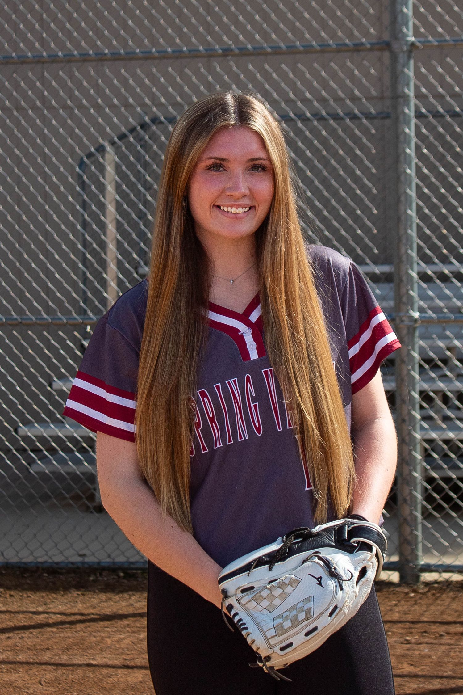 Long-haired softball player in Spring Valley jersey holds glove while posing on dirt field.
