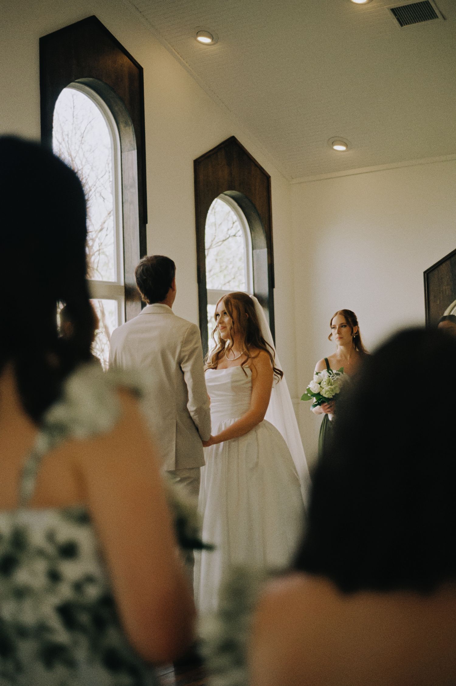 Bride in white gown smiling at groom during indoor wedding ceremony with arched windows and bridesmaid holding bouquet.