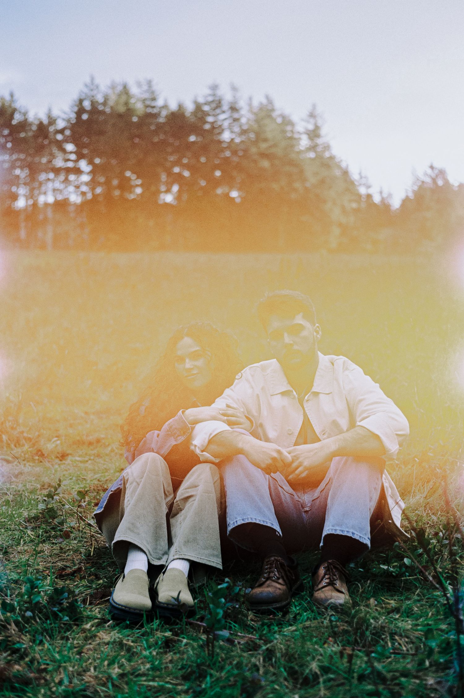 Couple sitting together on green grass with warm golden lens flare, trees in background, casual outdoor portrait.