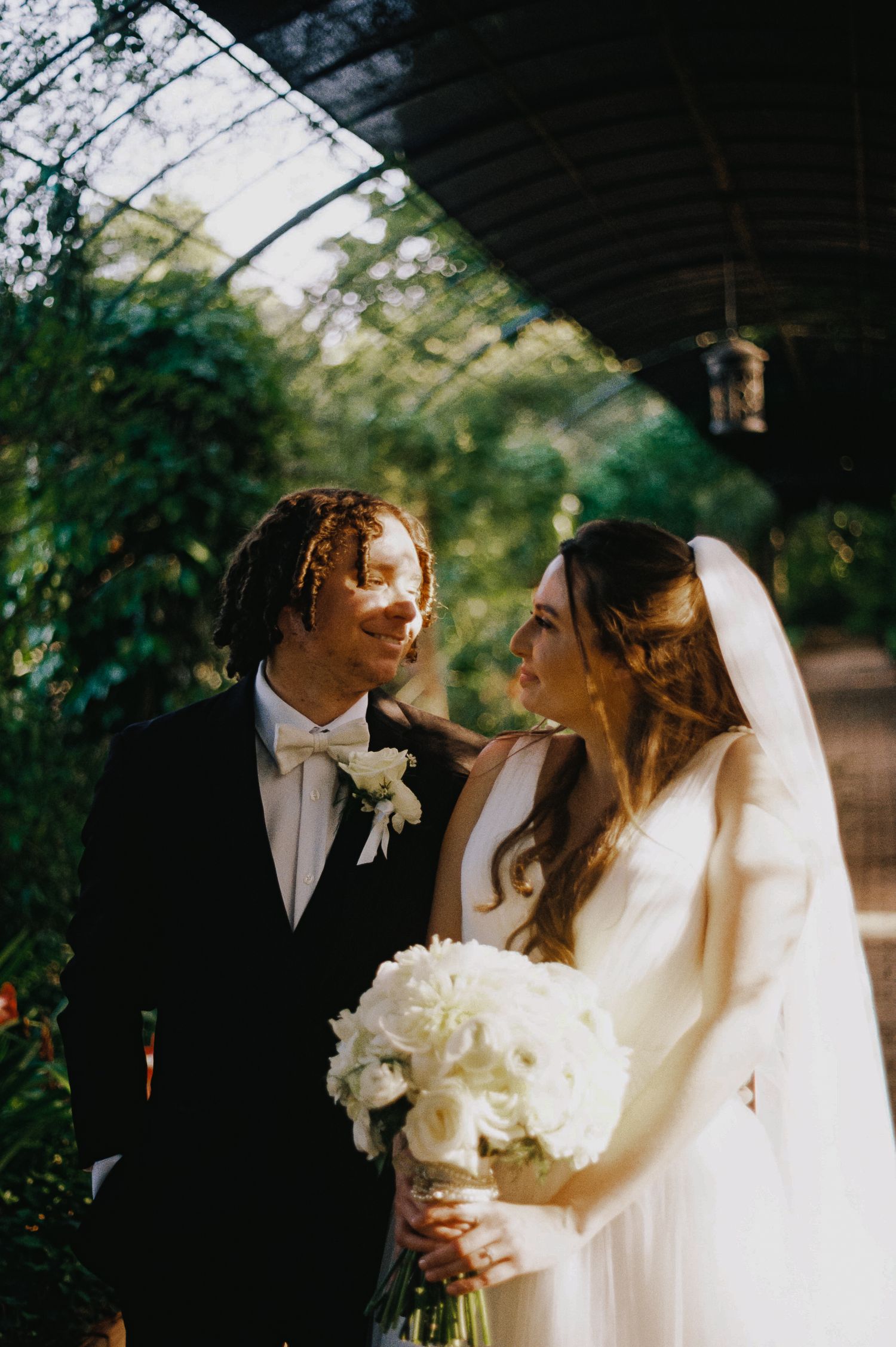 A bride and groom share a romantic moment outdoors, she holds white flowers, wearing a veil and gown, he wears a black tuxedo.