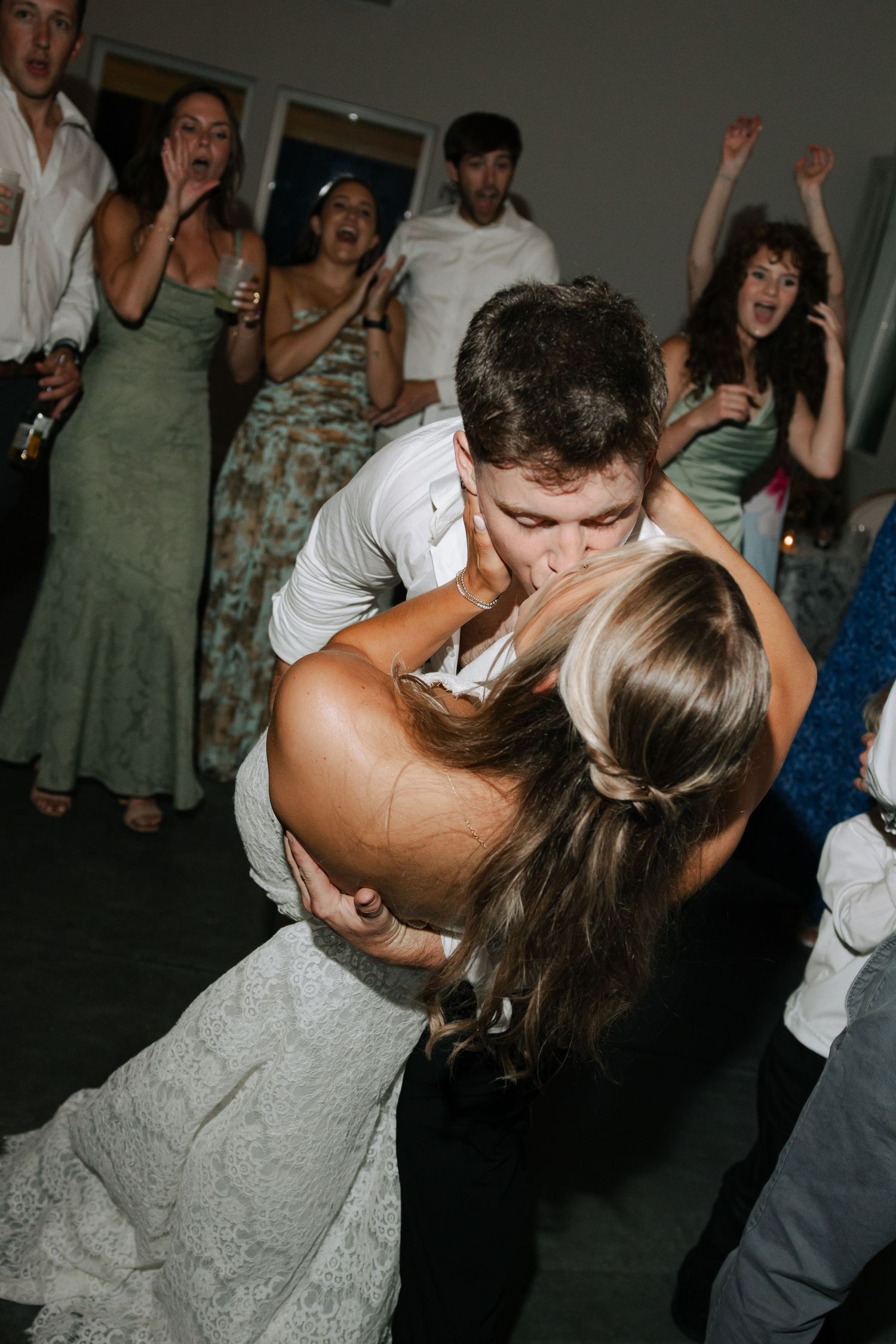 Bride and groom sharing a romantic dip kiss on the dance floor surrounded by cheering wedding guests.