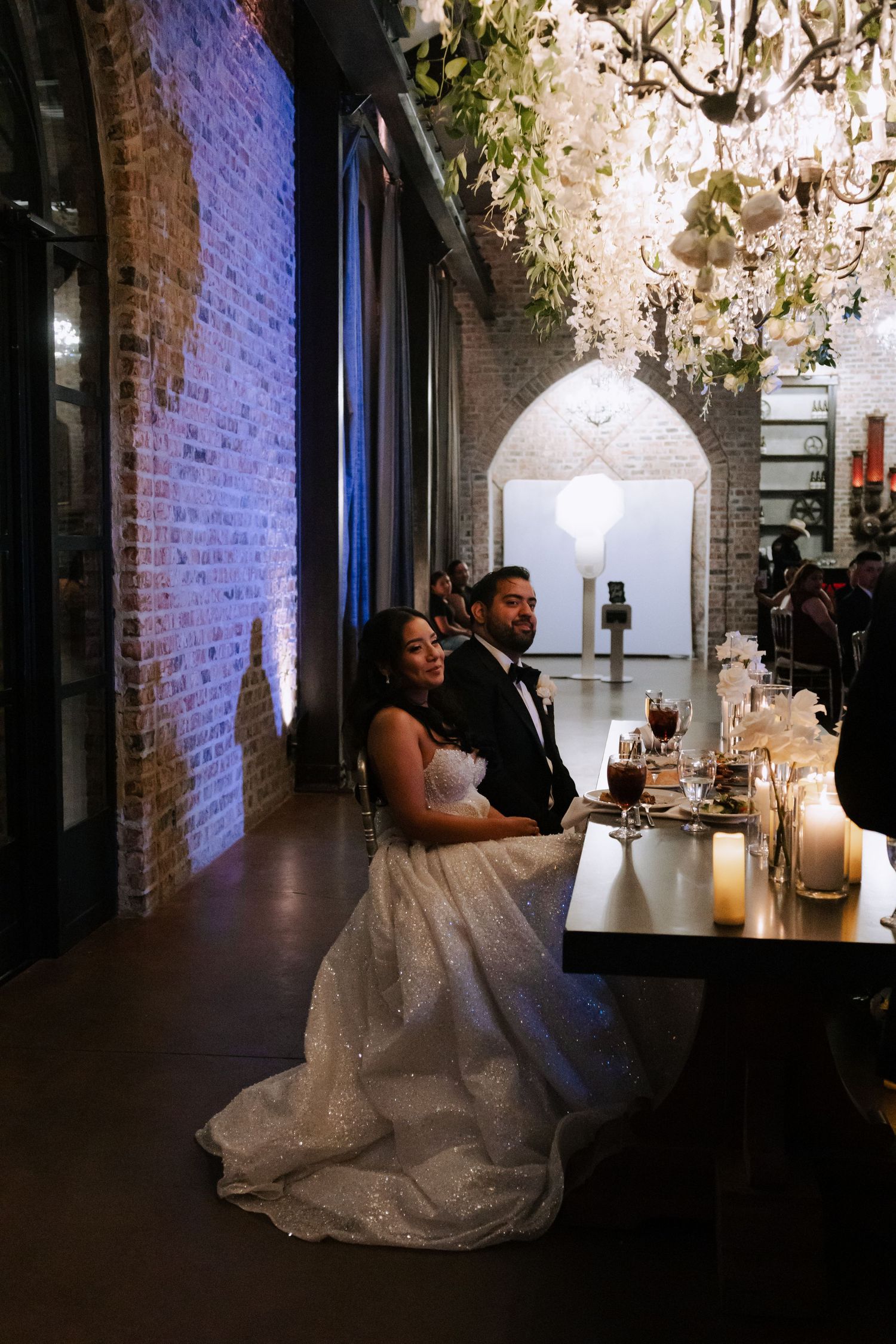 Bride and groom seated at their sweetheart table beneath a chandelier with candles and floral decor in an elegant brick venue.