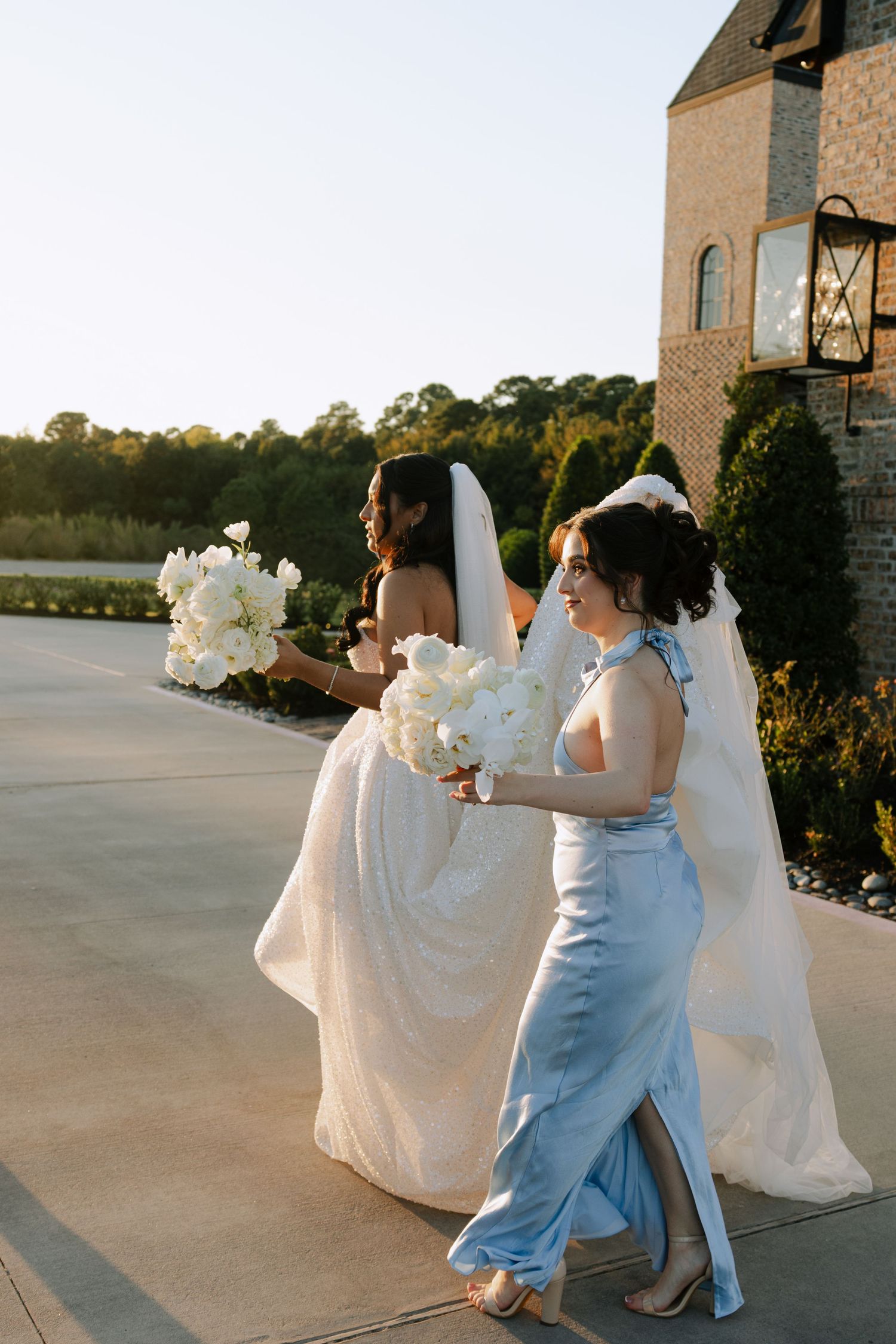 Bride in sequined gown and bridesmaid in blue dress walk outdoors holding white floral bouquets at golden hour.
