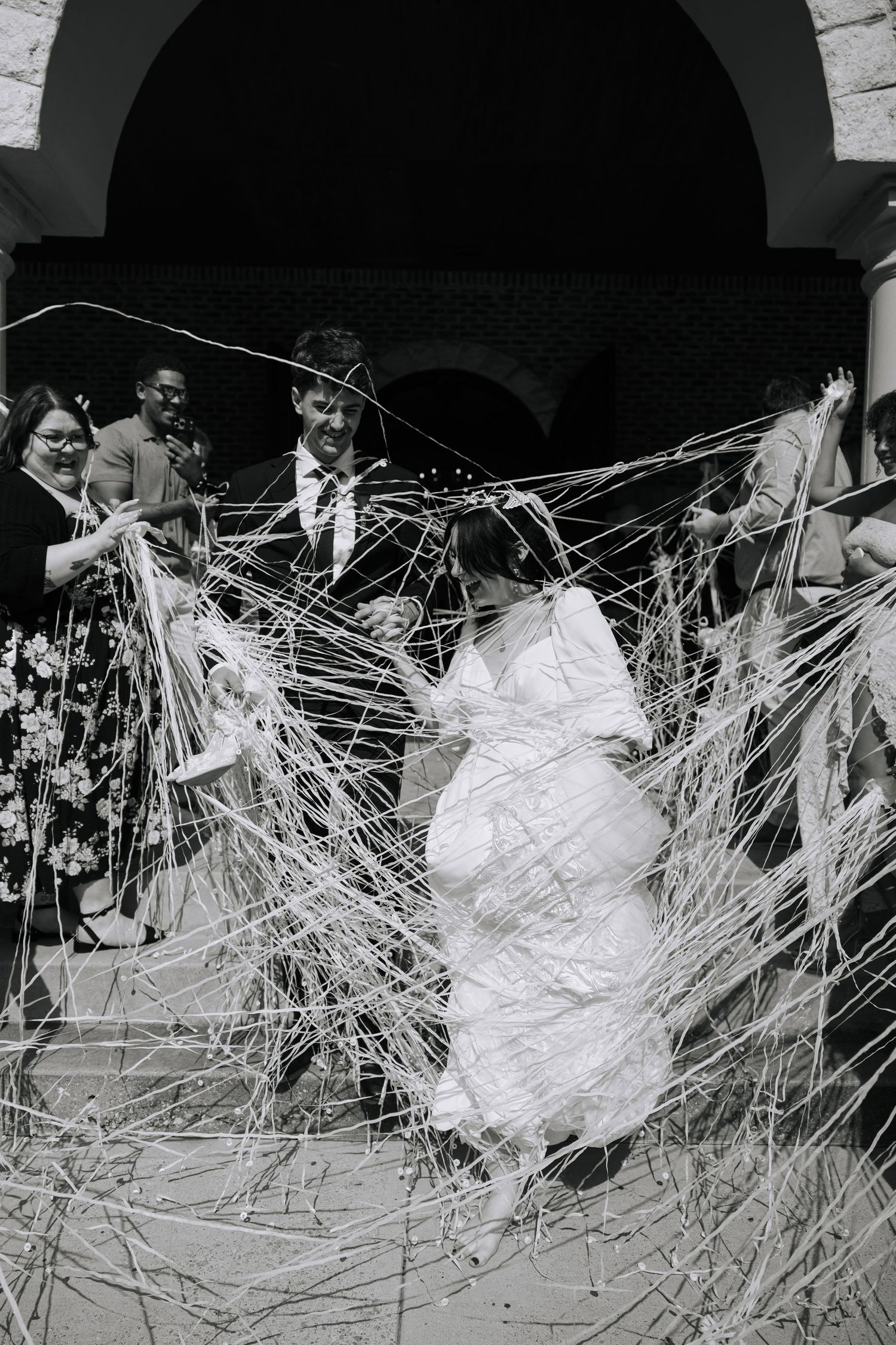 Bride and groom exit church through string confetti streamers, surrounded by cheering guests, in black and white wedding photo.