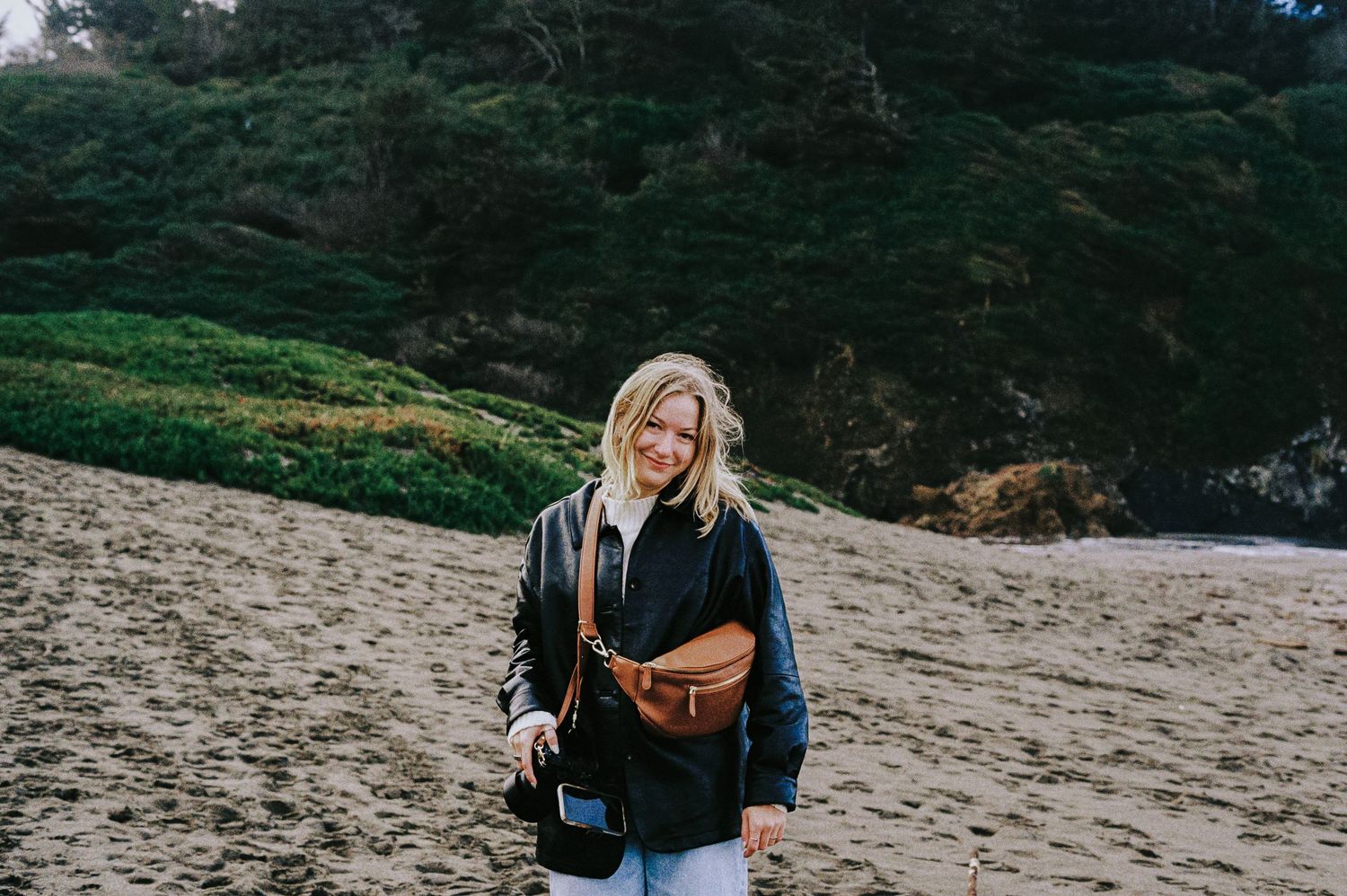 Smiling blonde woman in black jacket with brown crossbody bag standing on a sandy beach.