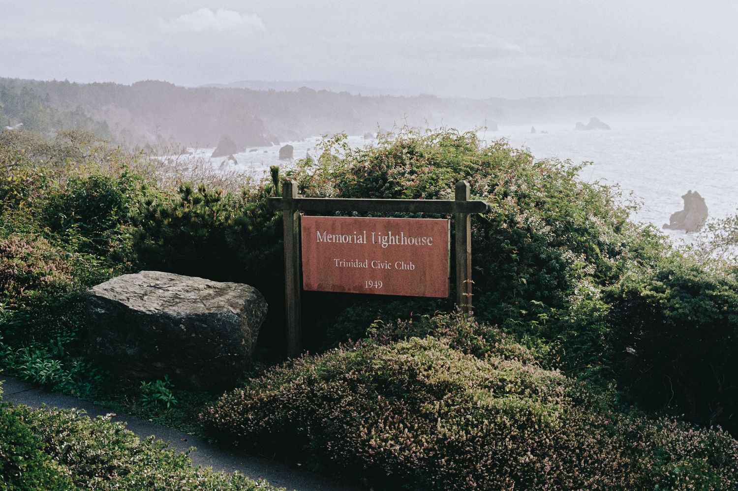 A wooden sign for Memorial Lighthouse surrounded by lush green shrubs with a misty coastal ocean view in the background.