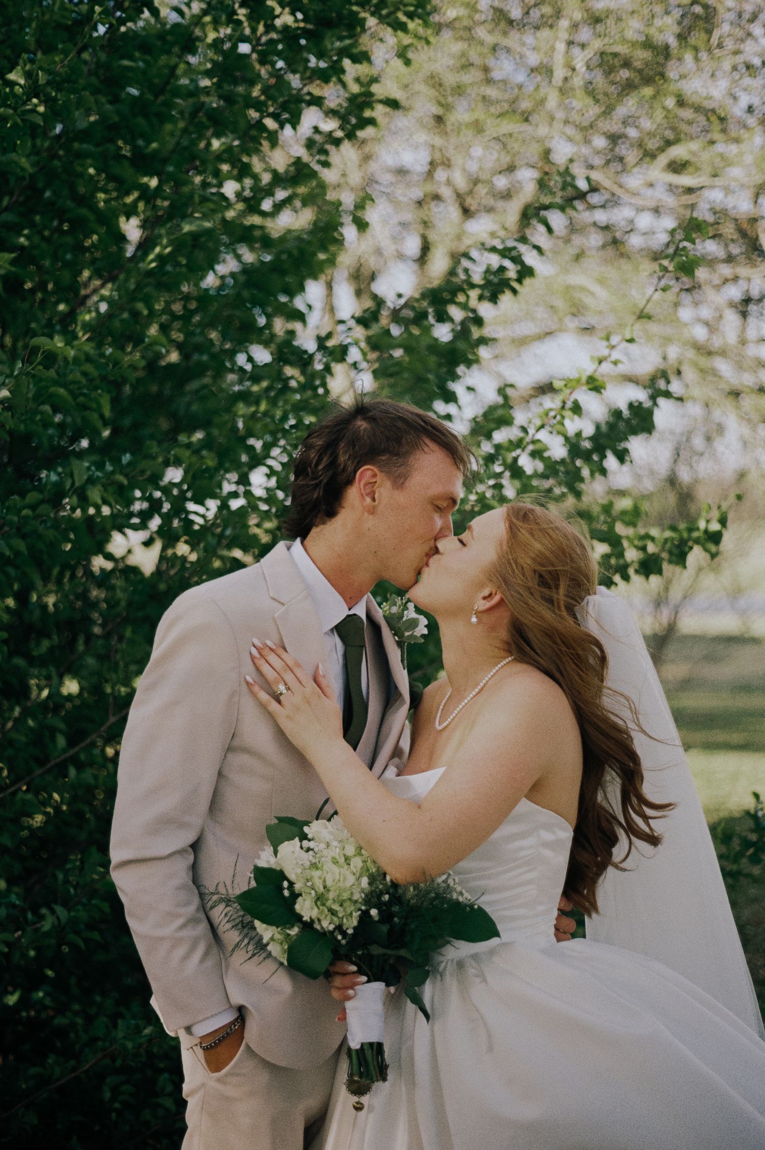 Bride and groom share a romantic kiss outdoors, surrounded by lush green trees, as she holds a white floral bouquet.