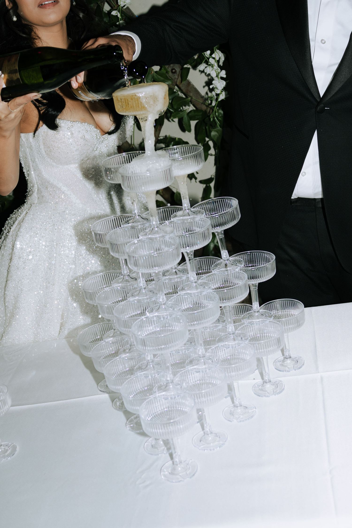 Bride and groom pouring champagne tower of crystal coupe glasses at elegant wedding reception.