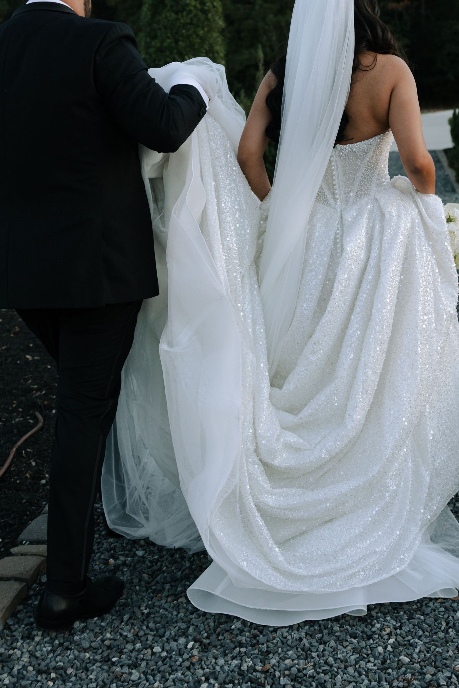 Back view of bride in sparkly sequined ballgown with flowing veil assisted by groom in black tuxedo outdoors.