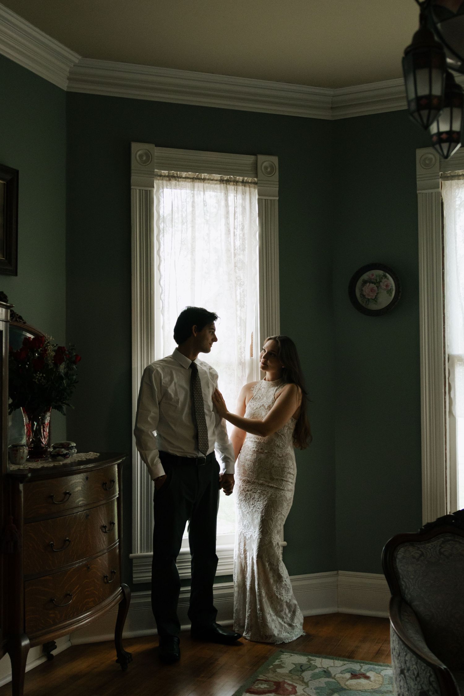 Bride and groom share a romantic moment by a window in a dimly lit vintage room with green walls and hardwood floors.