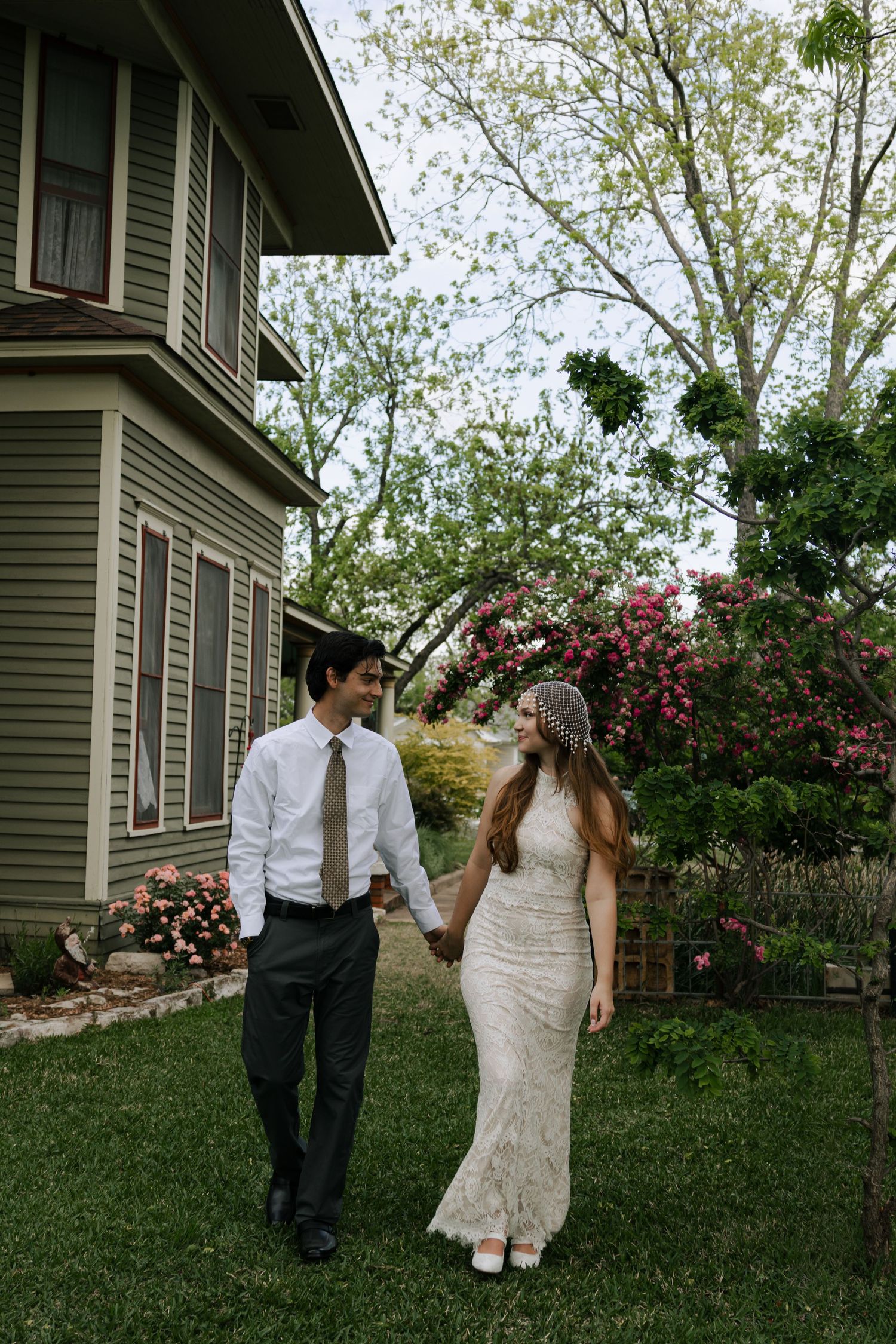 Bride and groom hold hands walking in lush backyard garden beside historic home on their wedding day.