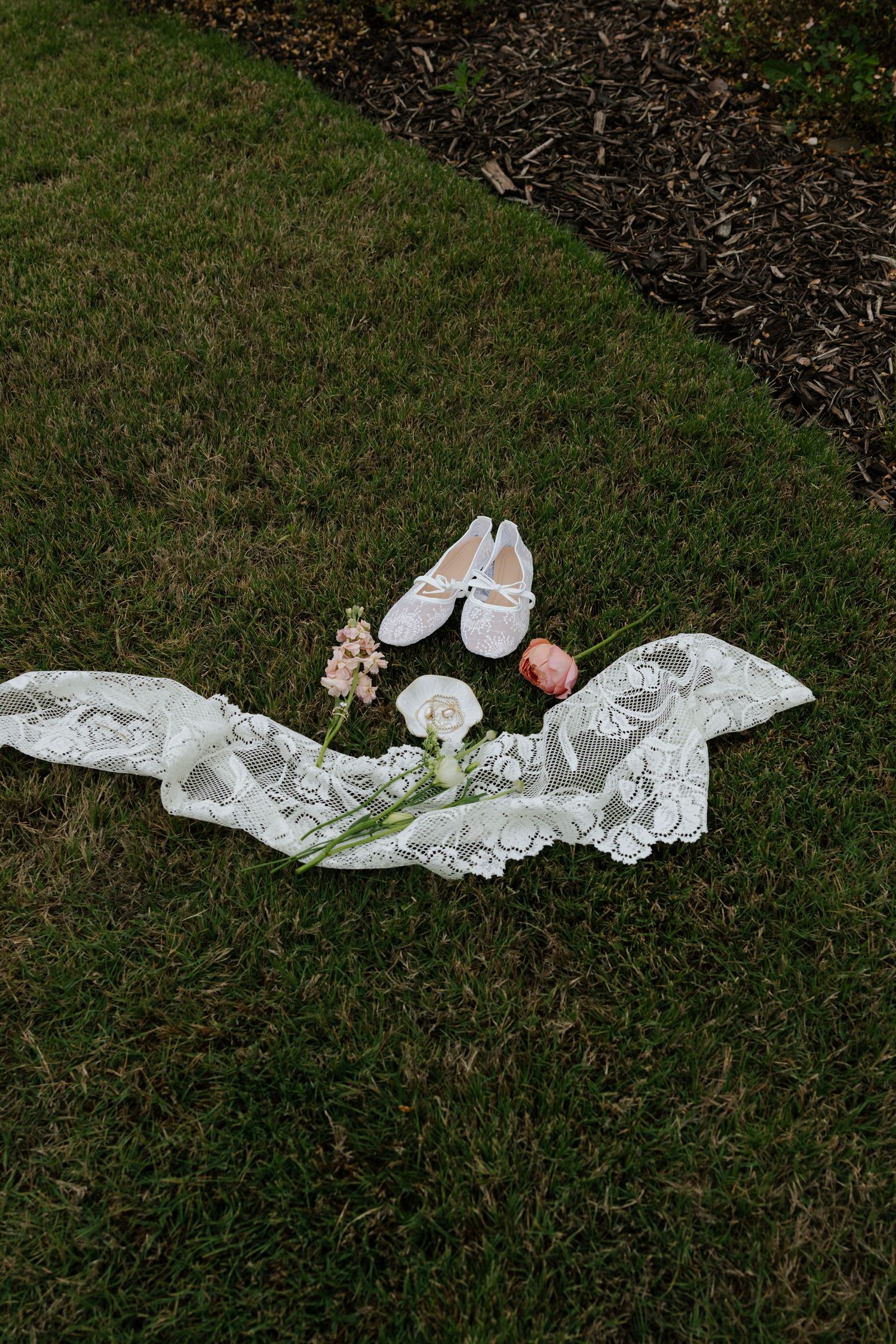 Bridal flat shoes, lace veil, and delicate flowers arranged on green grass for a wedding flat lay.
