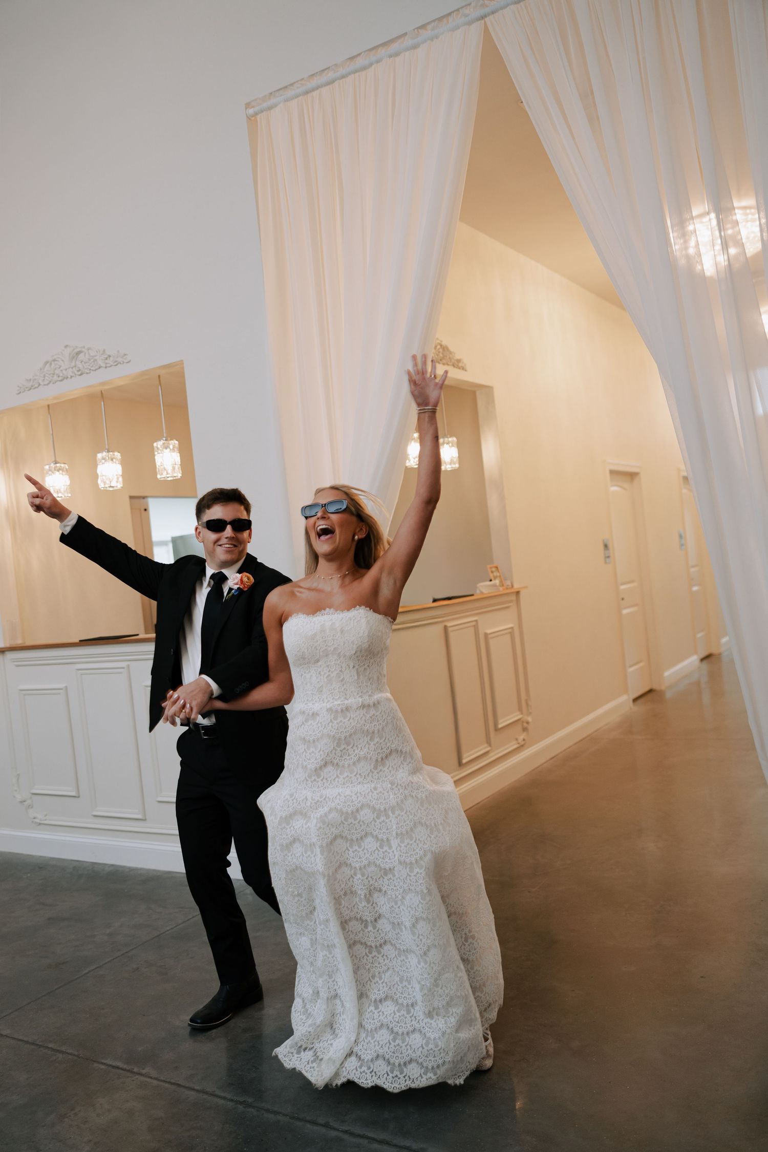 Bride and groom wearing sunglasses celebrate their grand reception entrance under draped white curtains.