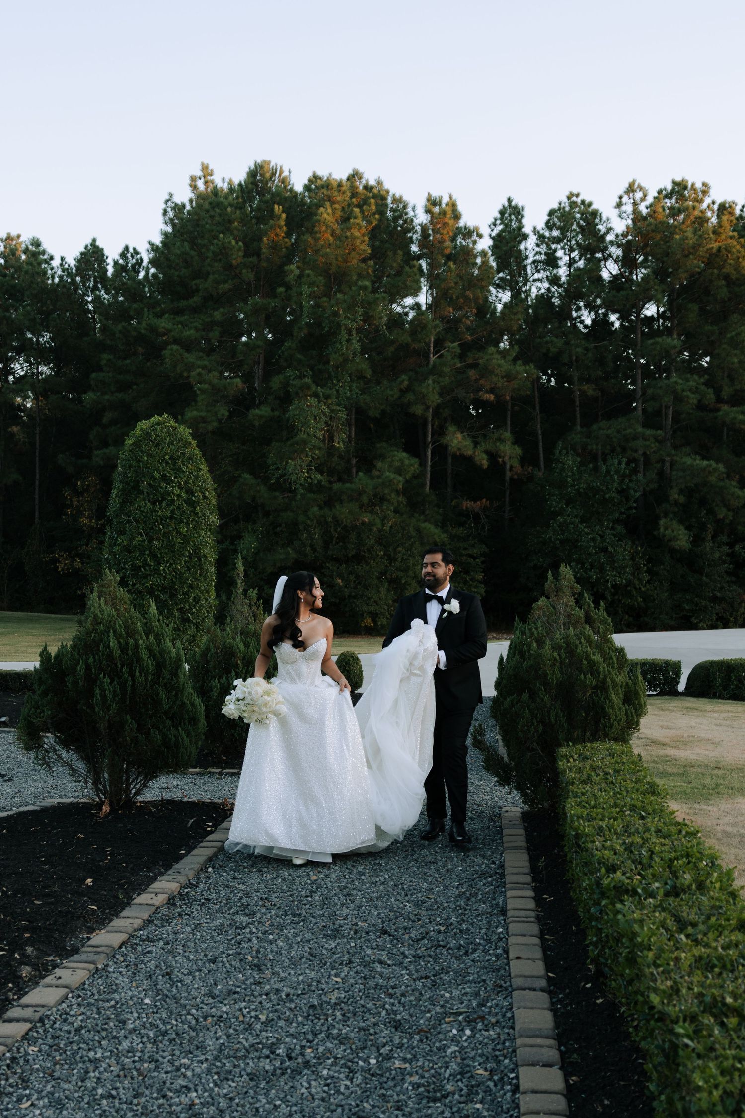 Elegant bride and groom walking through a manicured garden path at dusk surrounded by tall trees.