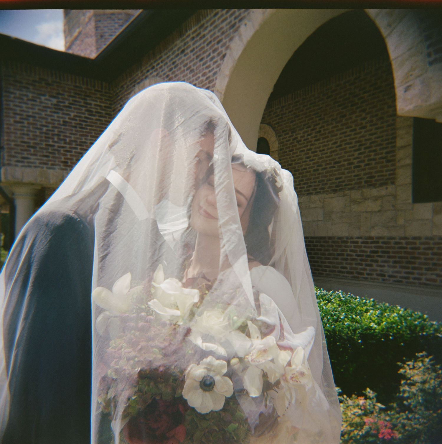 Bride and groom kissing under a flowing white wedding veil, holding a bouquet of white flowers outside a brick building.