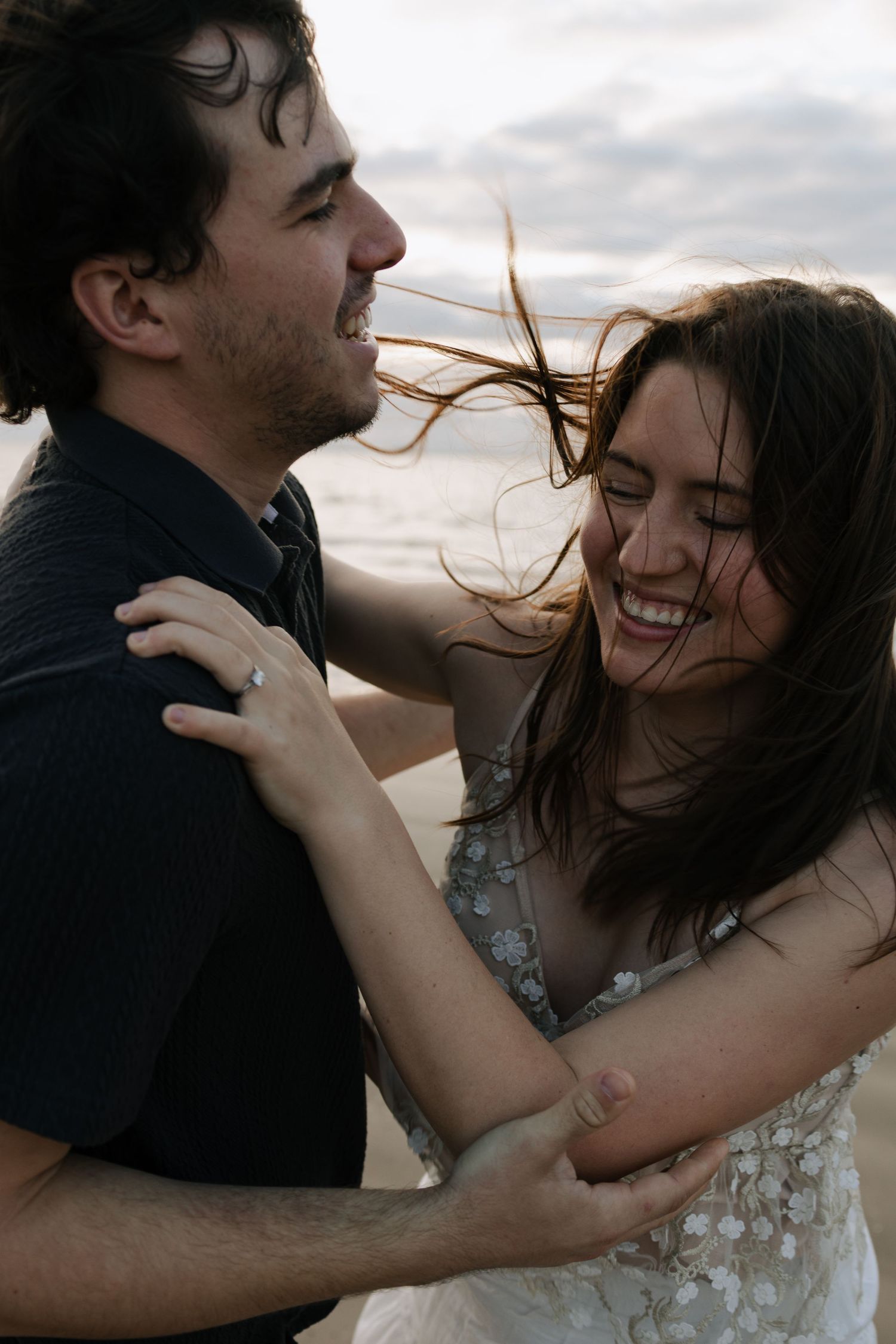 Laughing bride and groom embracing on a windy beach with hair blowing during romantic couples photo session.