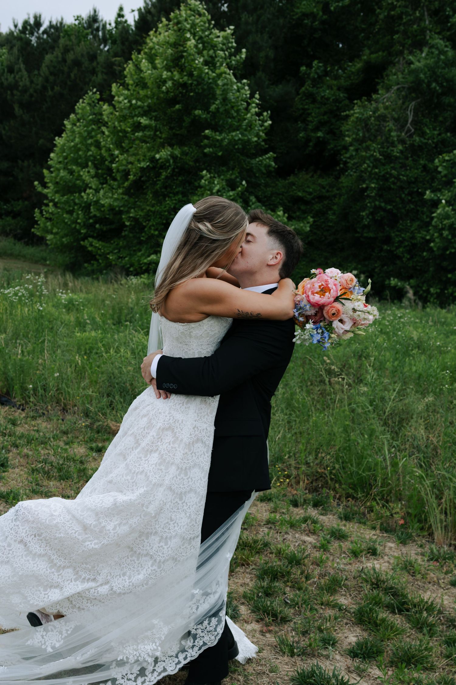 Bride and groom share a romantic kiss outdoors, with the bride holding a colorful bouquet in a lush green setting.