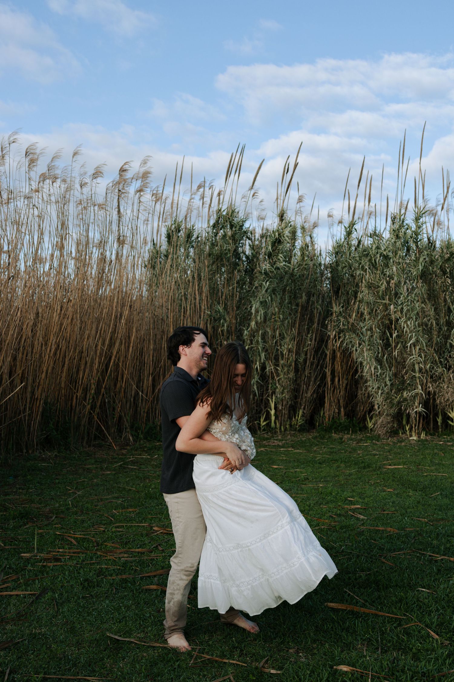 Happy couple dancing and laughing in a green field with tall golden reeds in the background.