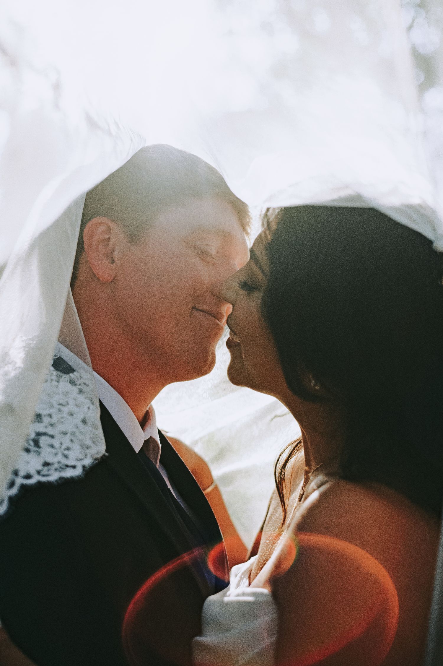 Bride and groom share a romantic moment under a lace wedding veil, bathed in soft natural light.