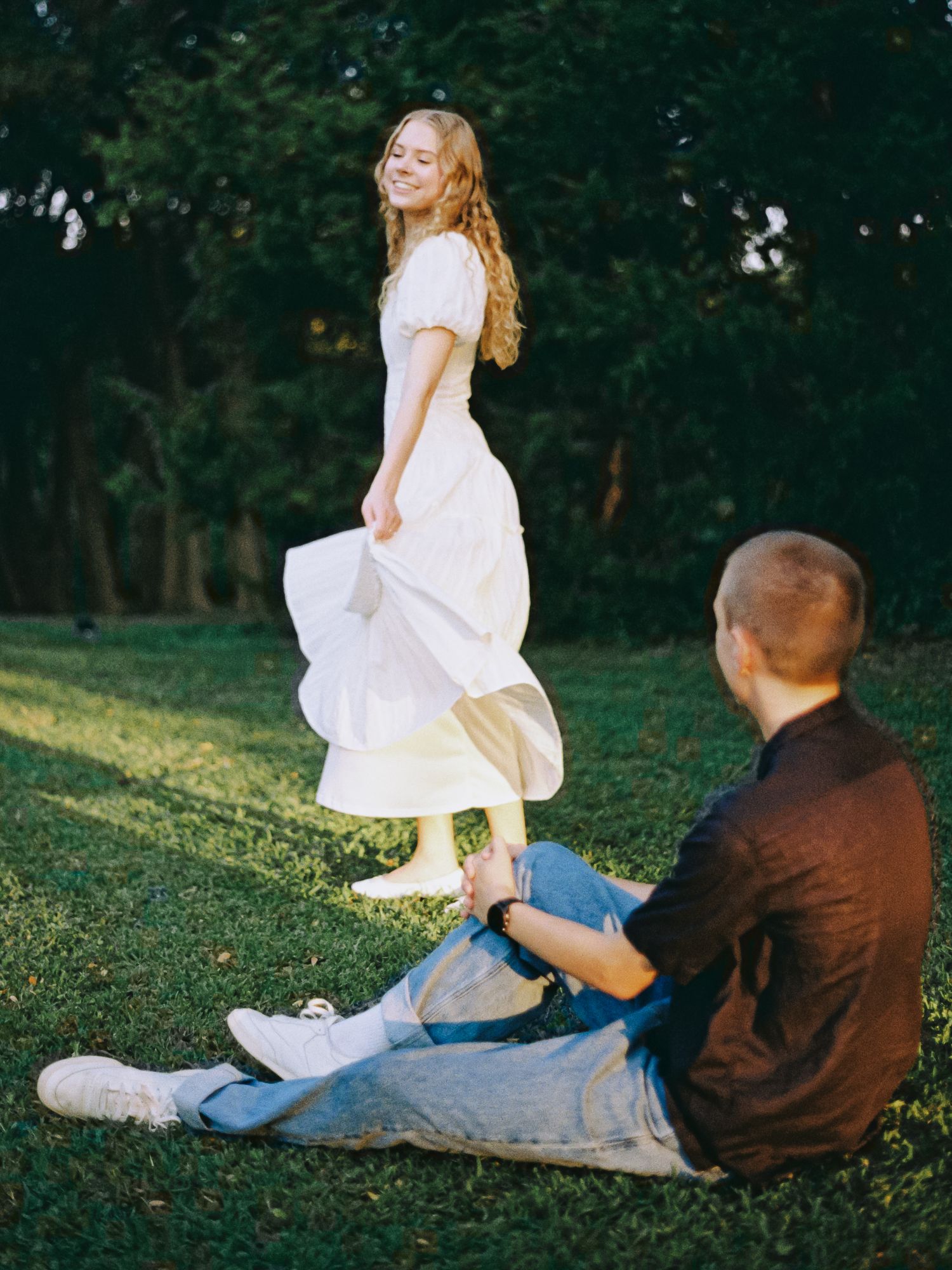 A smiling woman in a white dress twirls on green grass while a man sits watching her in a sunny park.