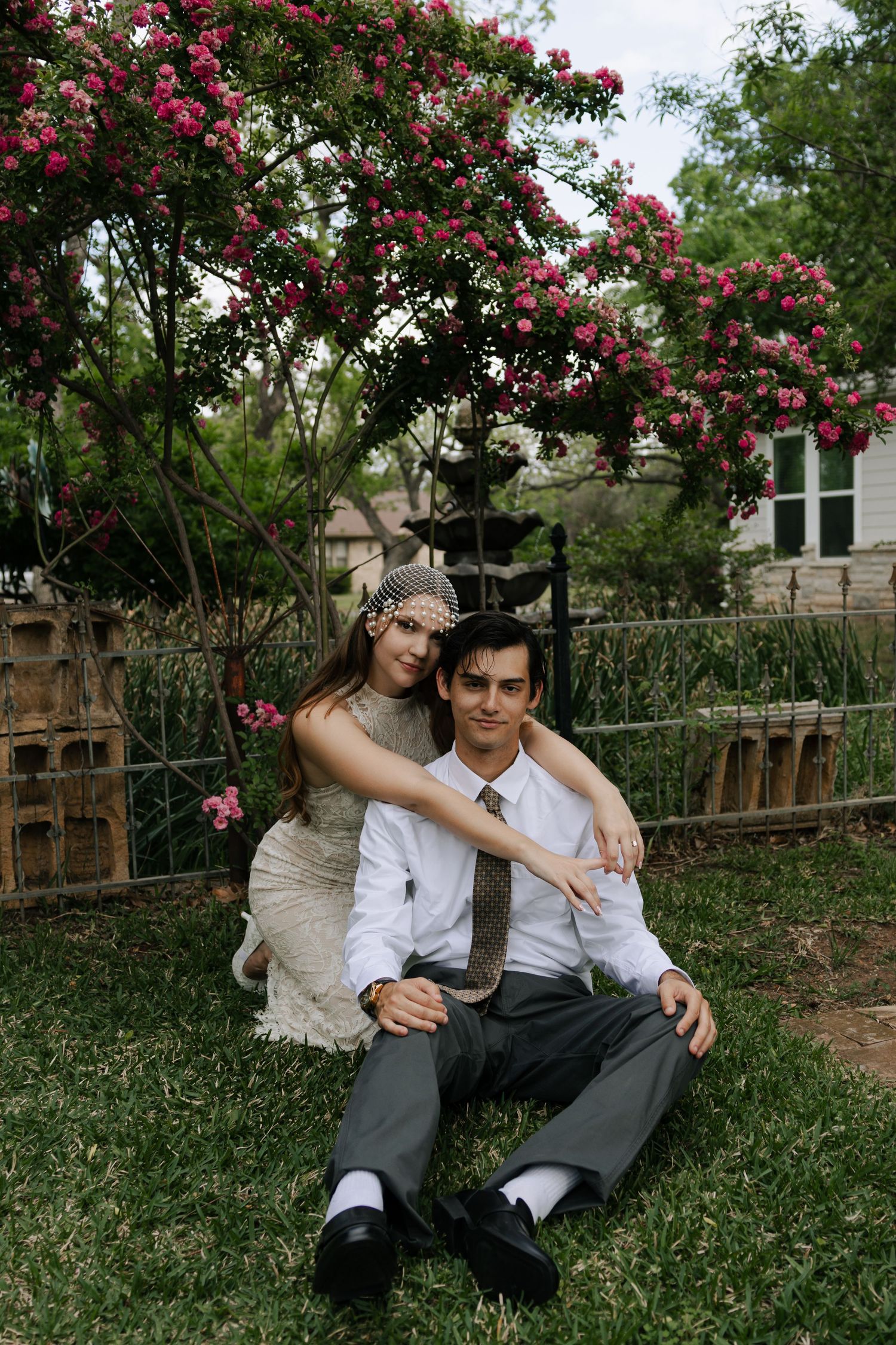Couple in vintage attire sitting on grass under blooming pink flower tree in a garden setting.