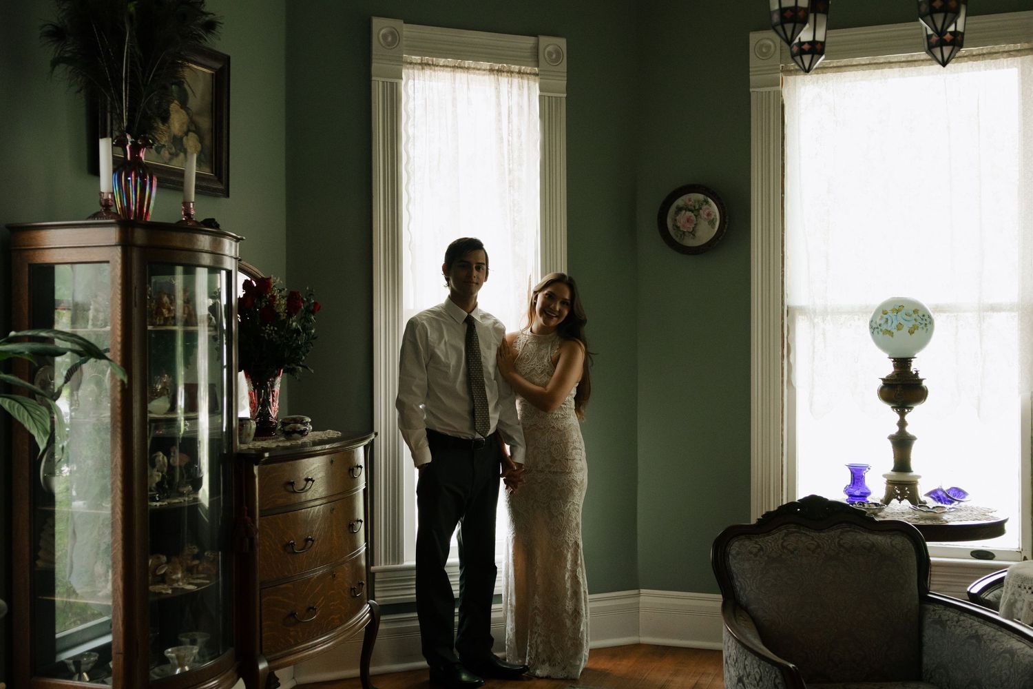 Couple in formal attire standing by bay window in elegant vintage room with antique furniture and green walls.