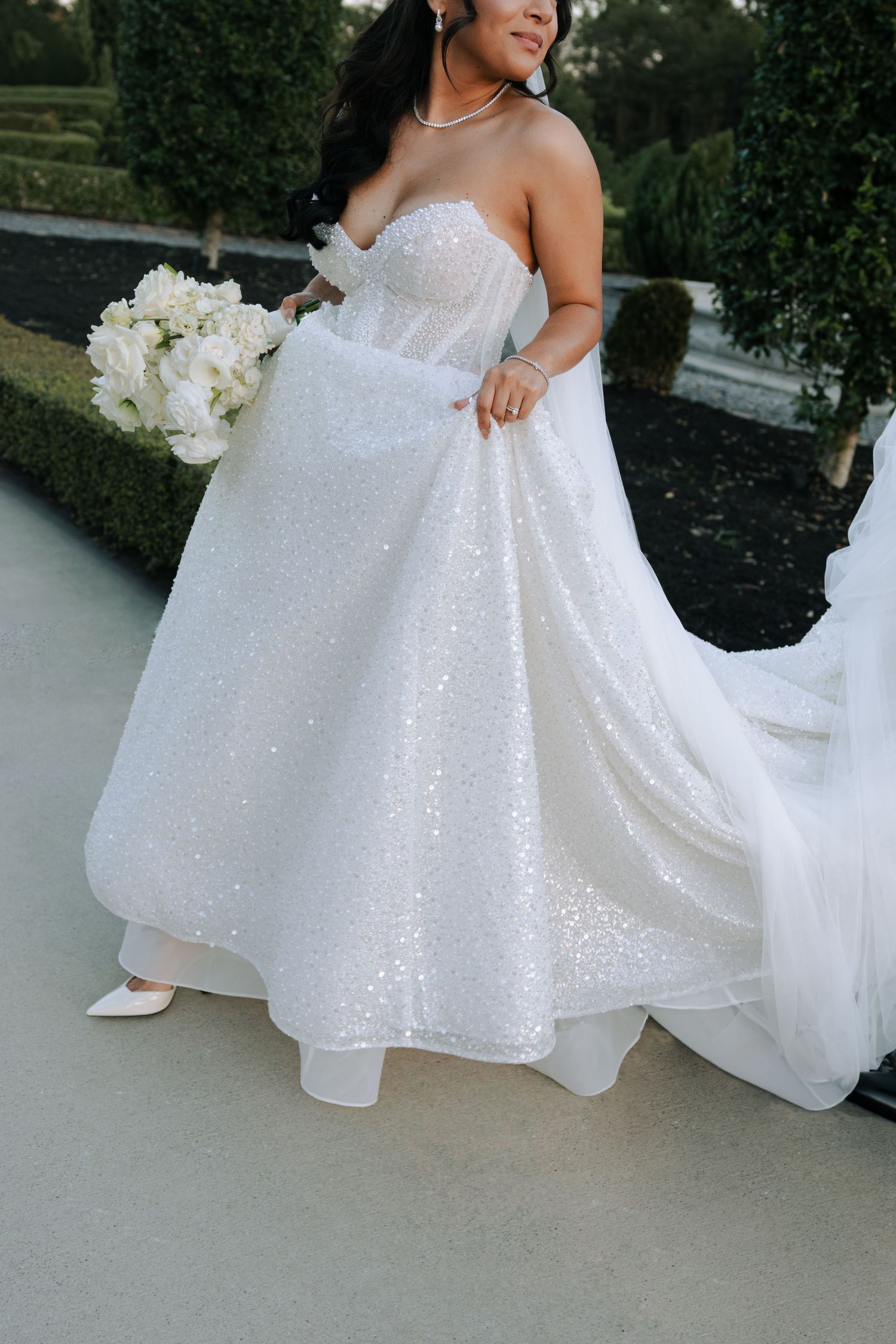 Close-up of bride in sequined strapless white ballgown holding white floral bouquet at outdoor wedding.