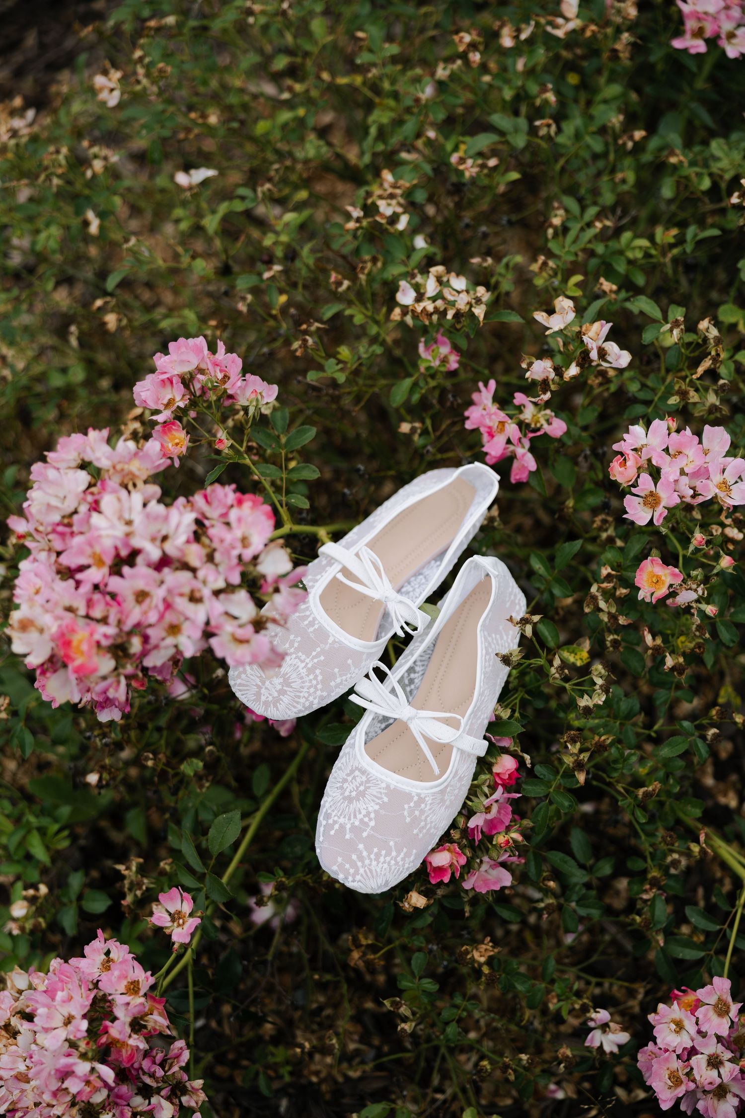 White lace bridal flats displayed among pink roses and green foliage in a garden setting.